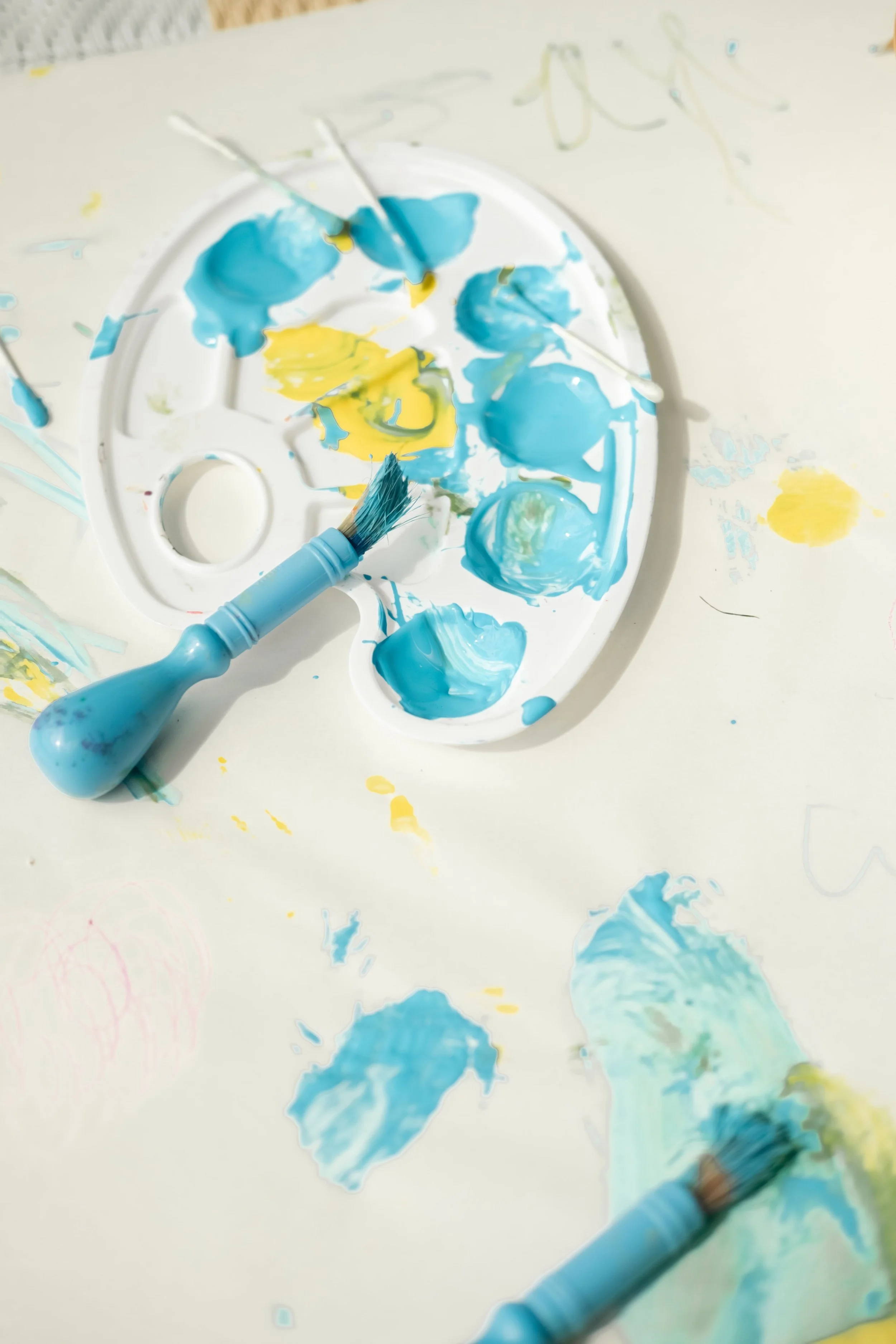 A white circular paint palette with blue and yellow paint blobs and two paintbrushes lying on it, surrounded by splatters and smudges of blue and yellow paint on a white surface.