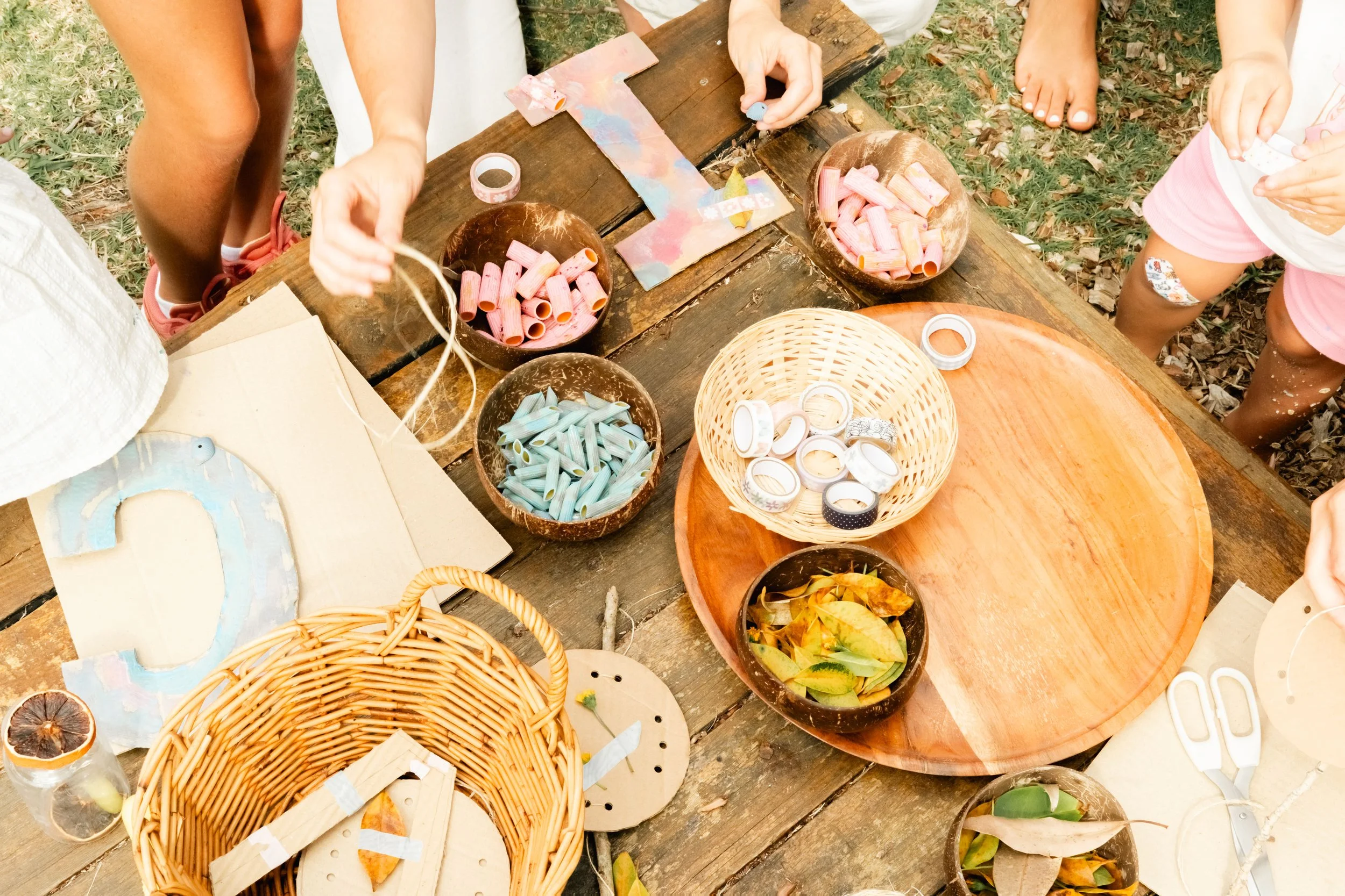 Children creating crafts with washi tape, paper, and other materials on a wooden table outdoors.