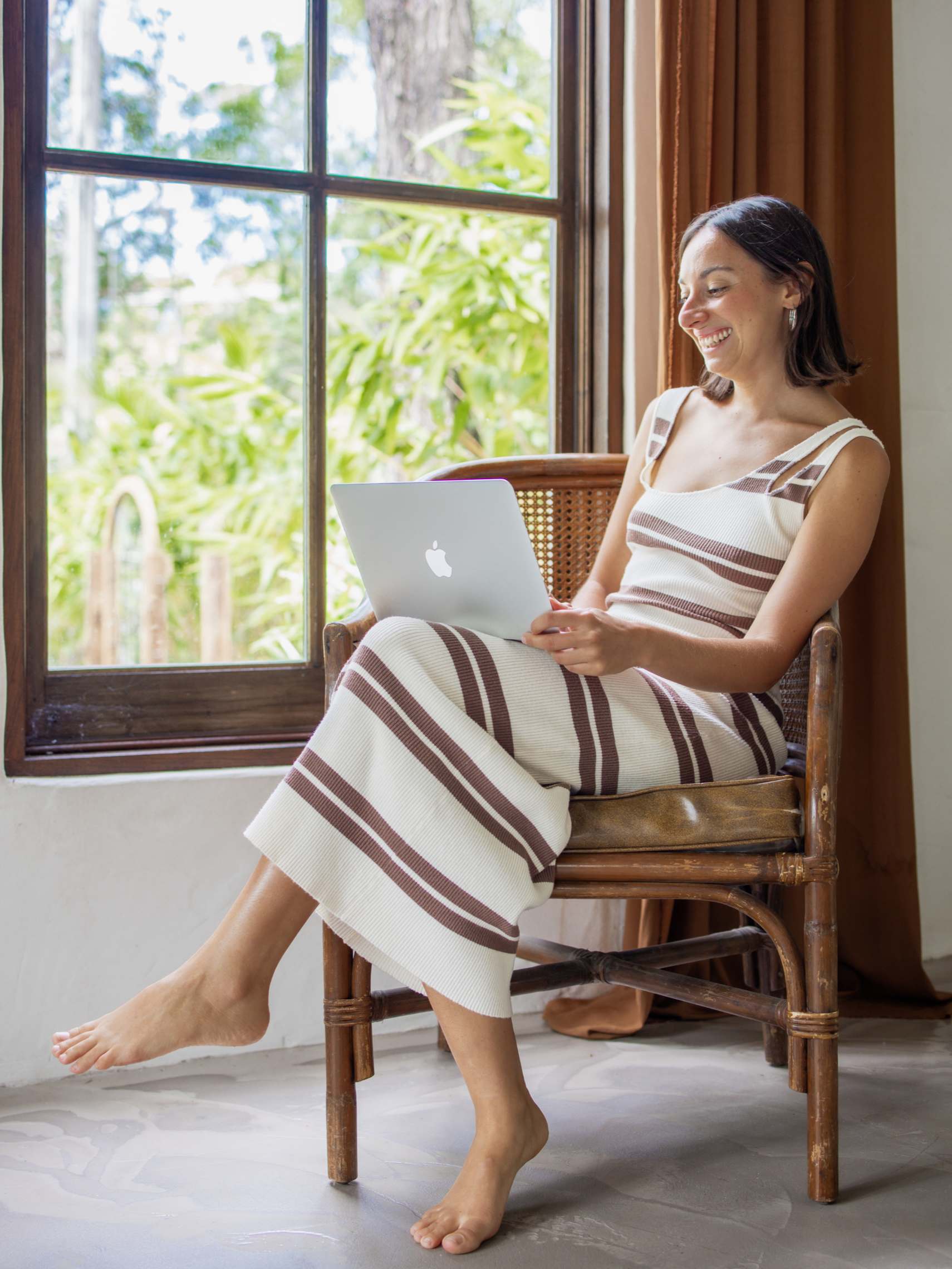 Woman sitting on a wooden chair near a window, smiling and working on a laptop.