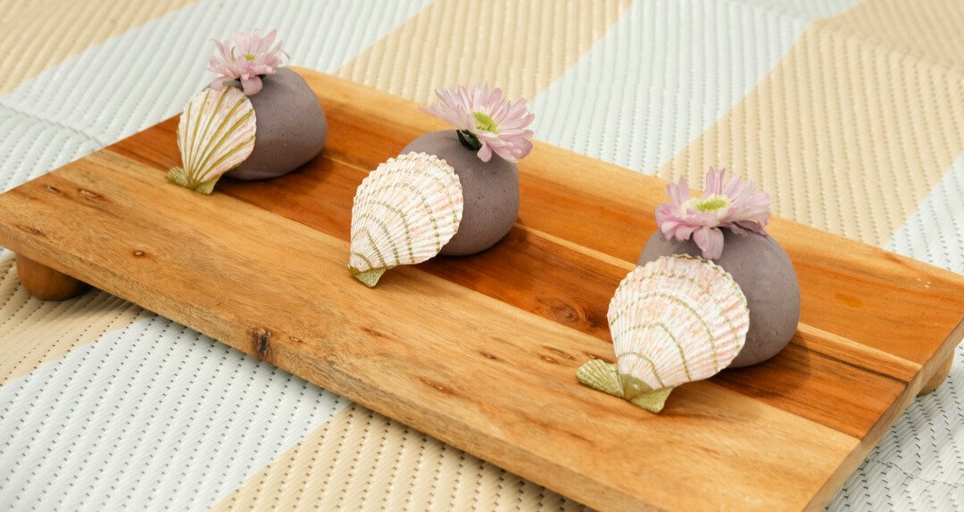 Decorative wooden tray holding three small gray vases with pink flowers and seashells, placed on a light-colored, striped fabric surface.