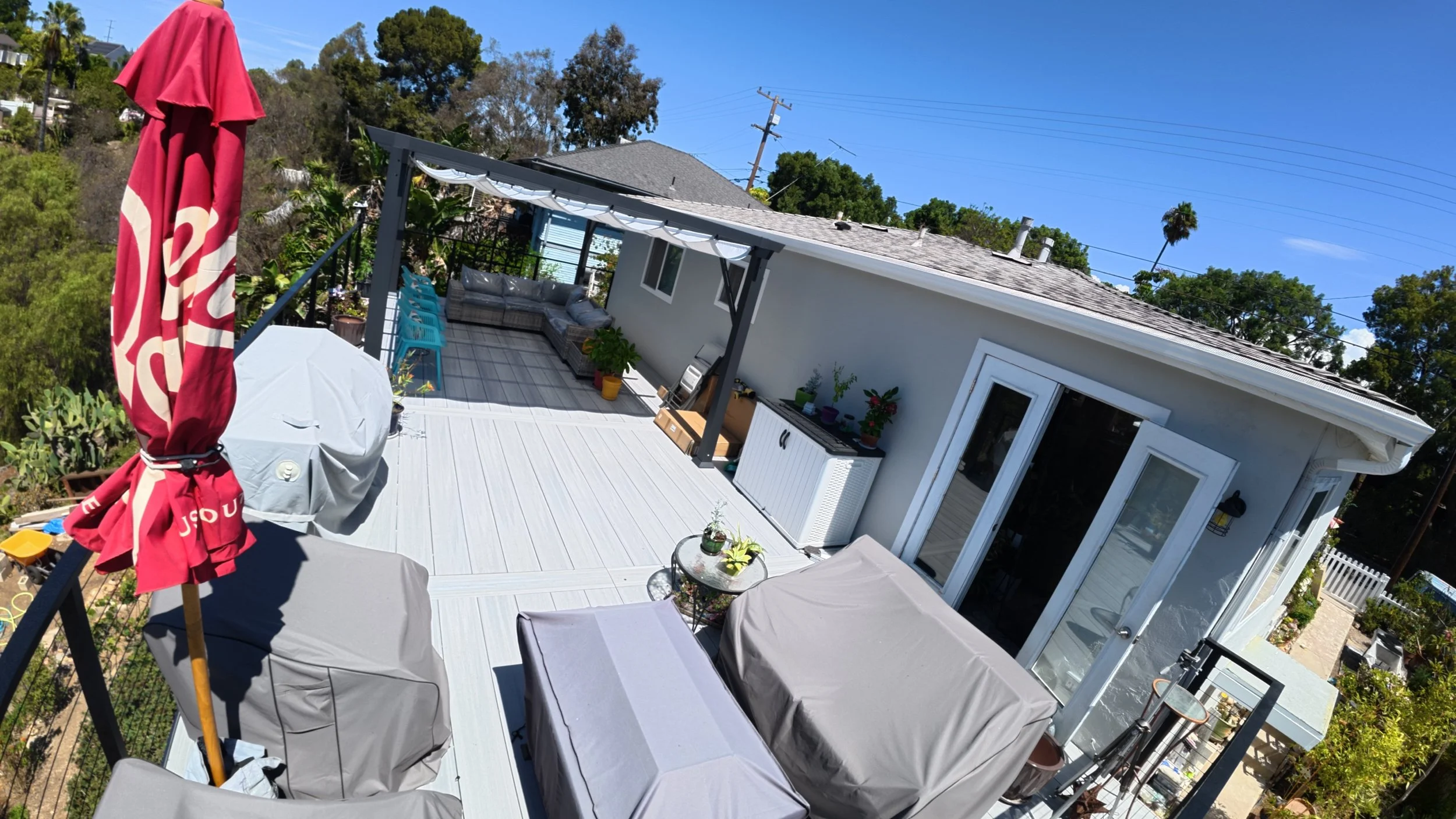 A backyard deck on a sunny day with outdoor furniture, a large red umbrella, and plants, attached to a house with white siding and a sliding glass door.