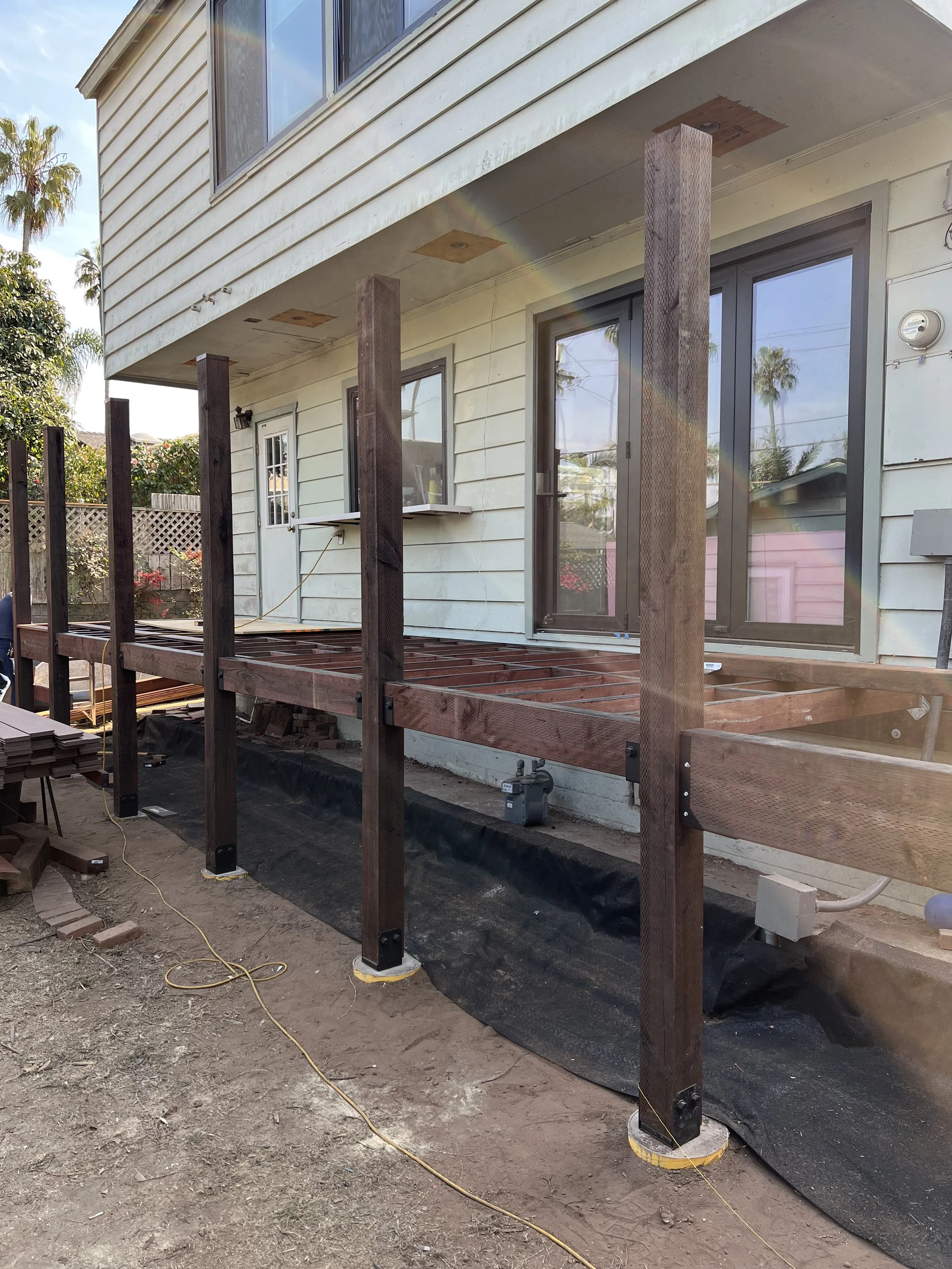 Backyard construction site with a wooden deck under construction. The deck is supported by vertical wooden posts anchored with metal brackets, and a black foundation beneath. The house has white siding, sliding glass doors, and a small window, with some construction tools and materials scattered around the dirt ground.