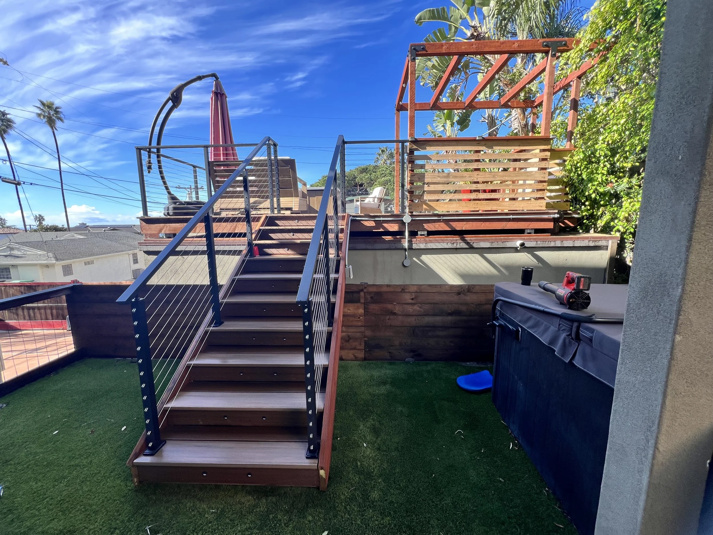 Backyard deck under construction with wooden stairs, metal railings, and a wooden frame structure, surrounded by green artificial grass and outdoor furniture.