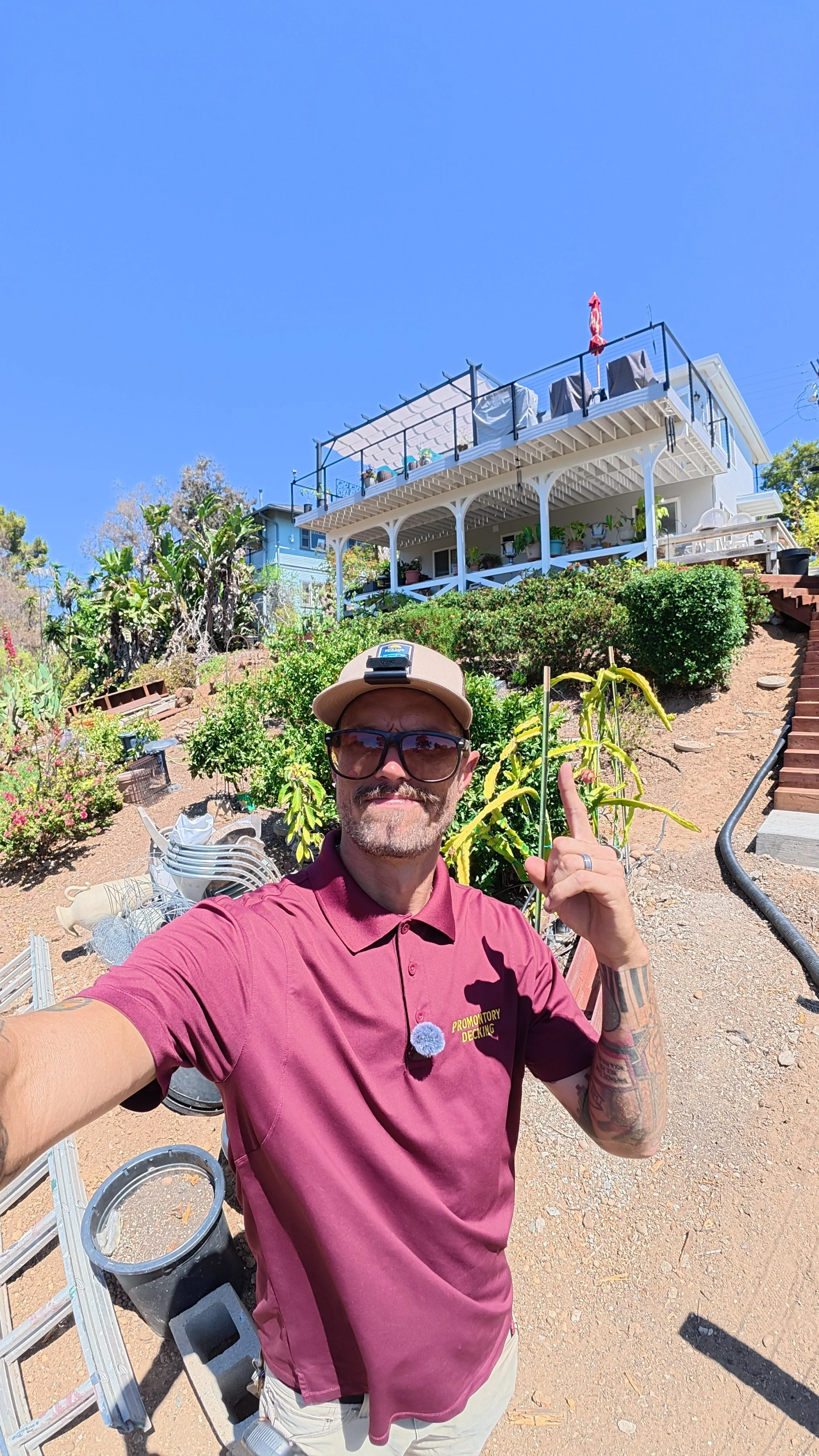 Man taking a selfie outdoors in front of a house with a terrace and climbing plants. He is wearing sunglasses, a beige cap, a maroon polo shirt with a logo, and has tattoos on his arm. The sky is clear and blue.
