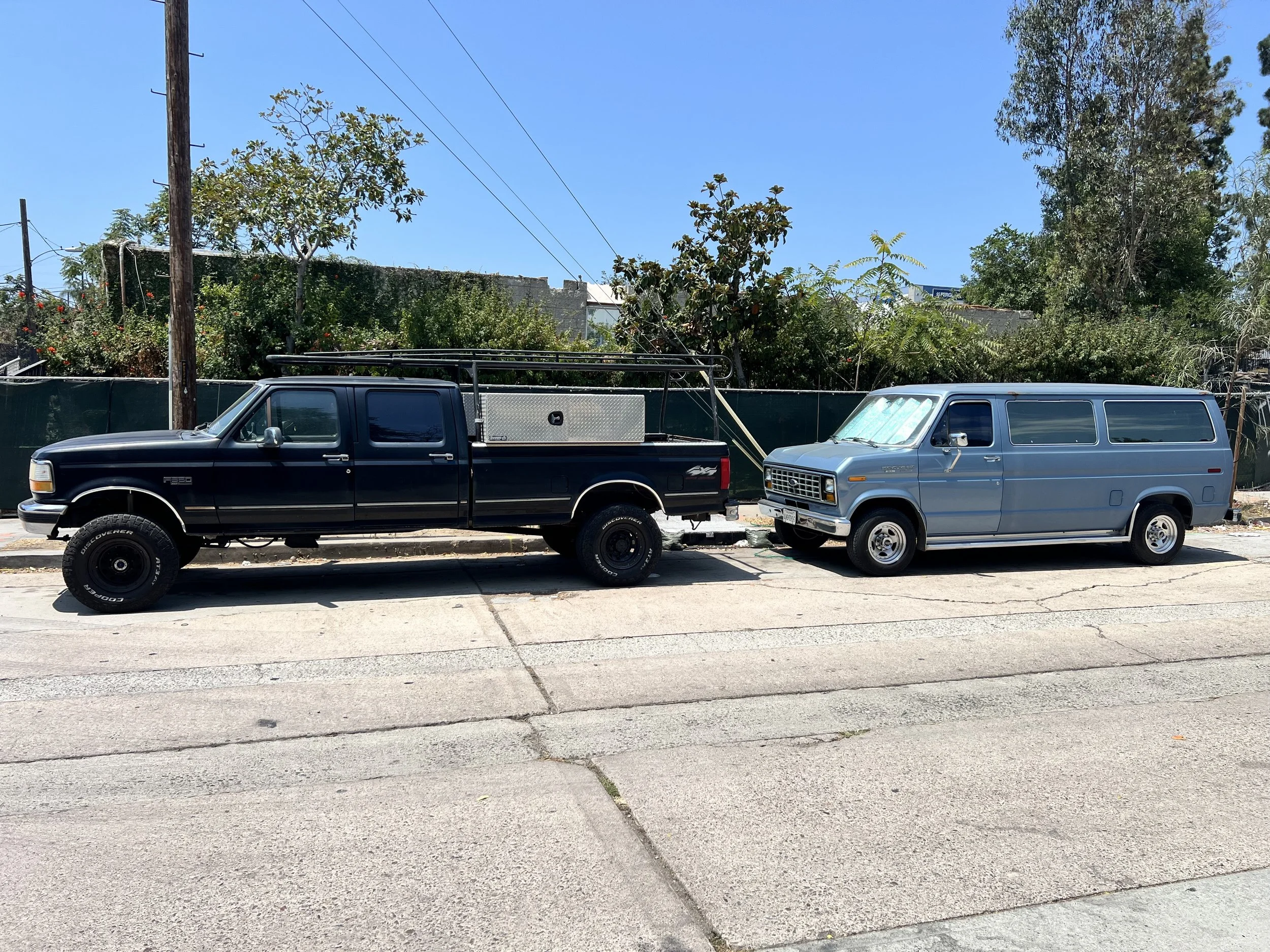 Two vehicles parked on a street in front of a fence with trees and power lines in the background, under a clear blue sky.