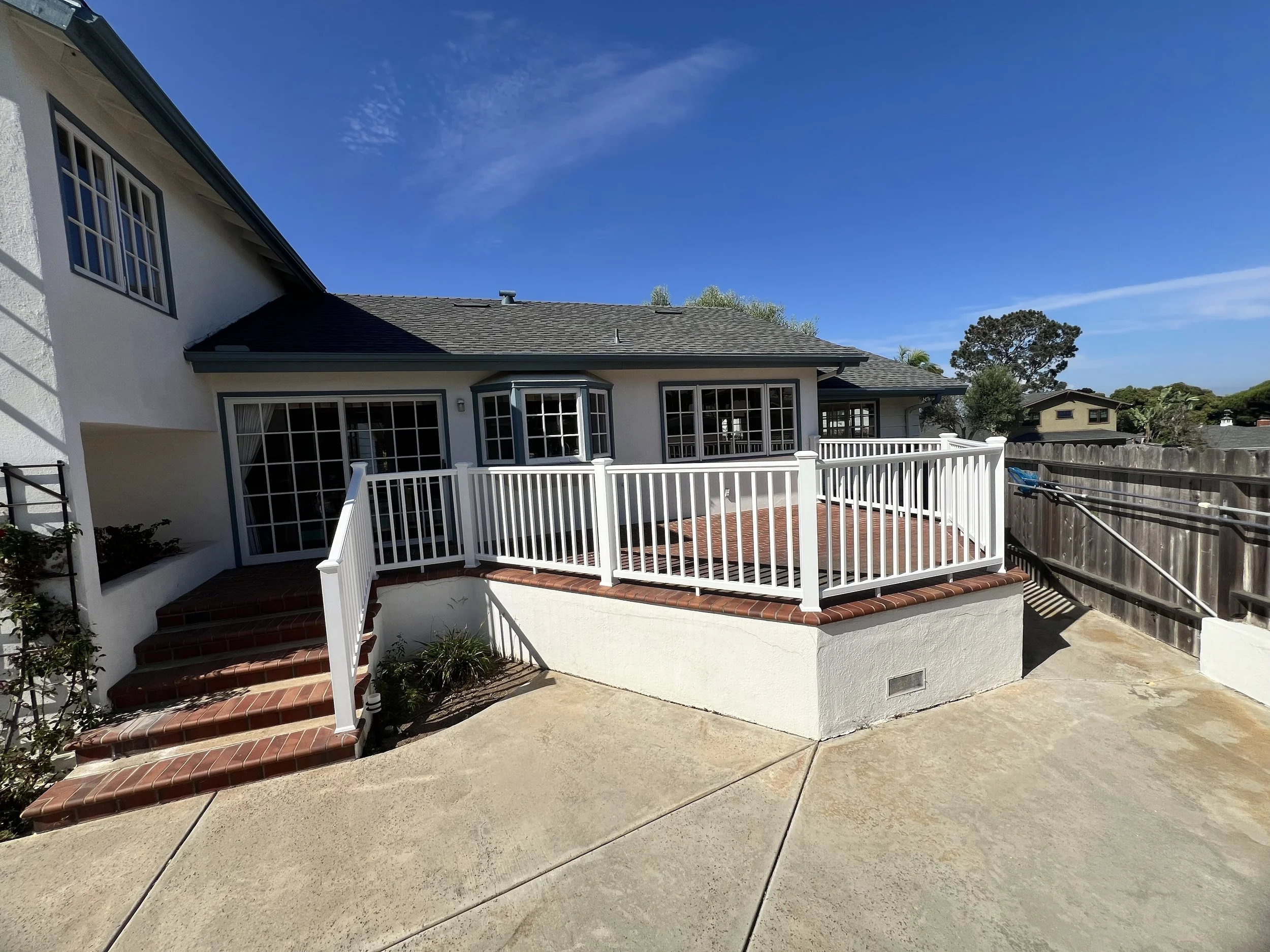 Backyard patio area with stairs, white railing, and a house with large windows and a shingled roof.