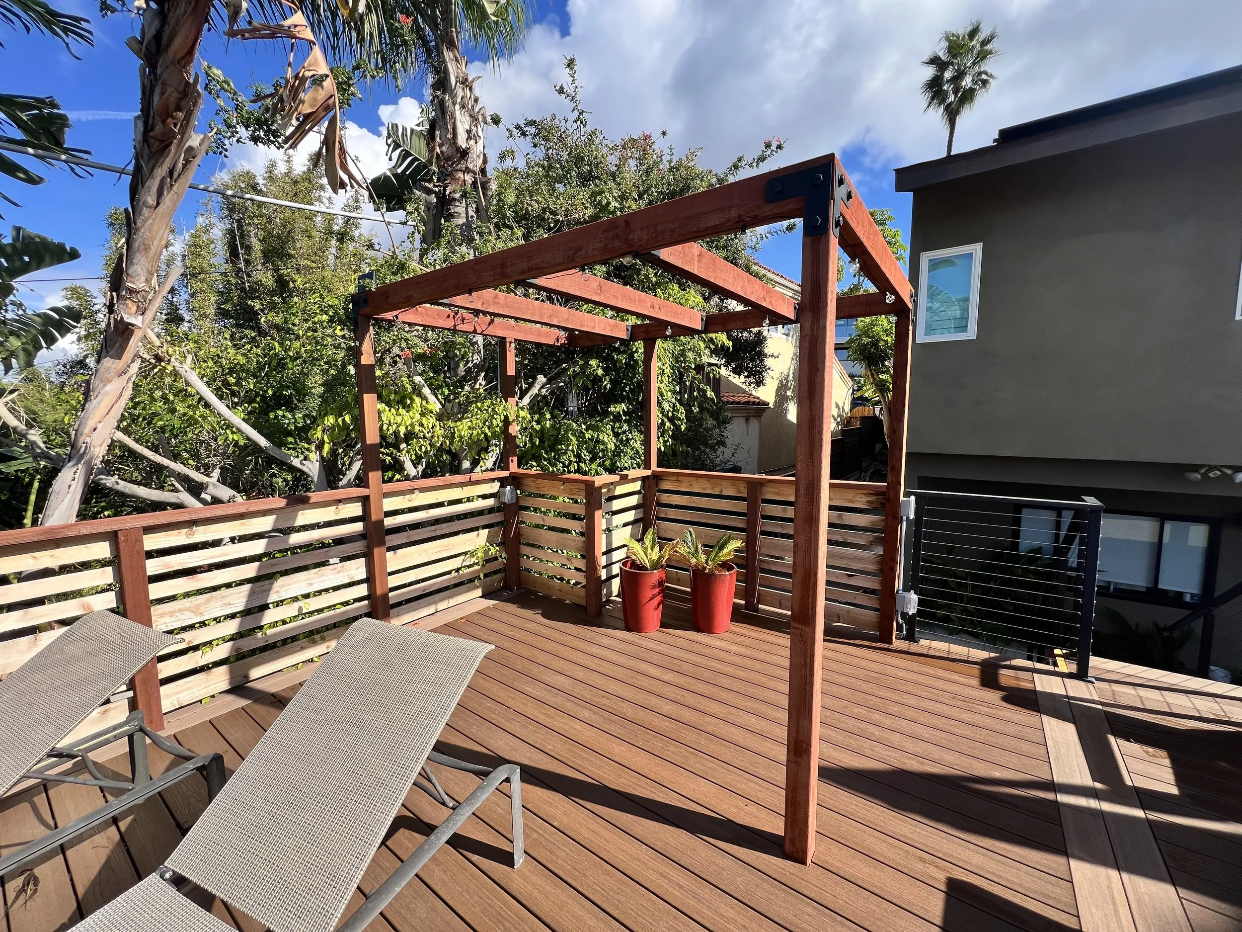 A wooden deck in a backyard with two reclined lounge chairs, a partially constructed wooden pergola, two red potted plants, and lush green trees and shrubs. A neighboring house is visible in the background under a partly cloudy sky.