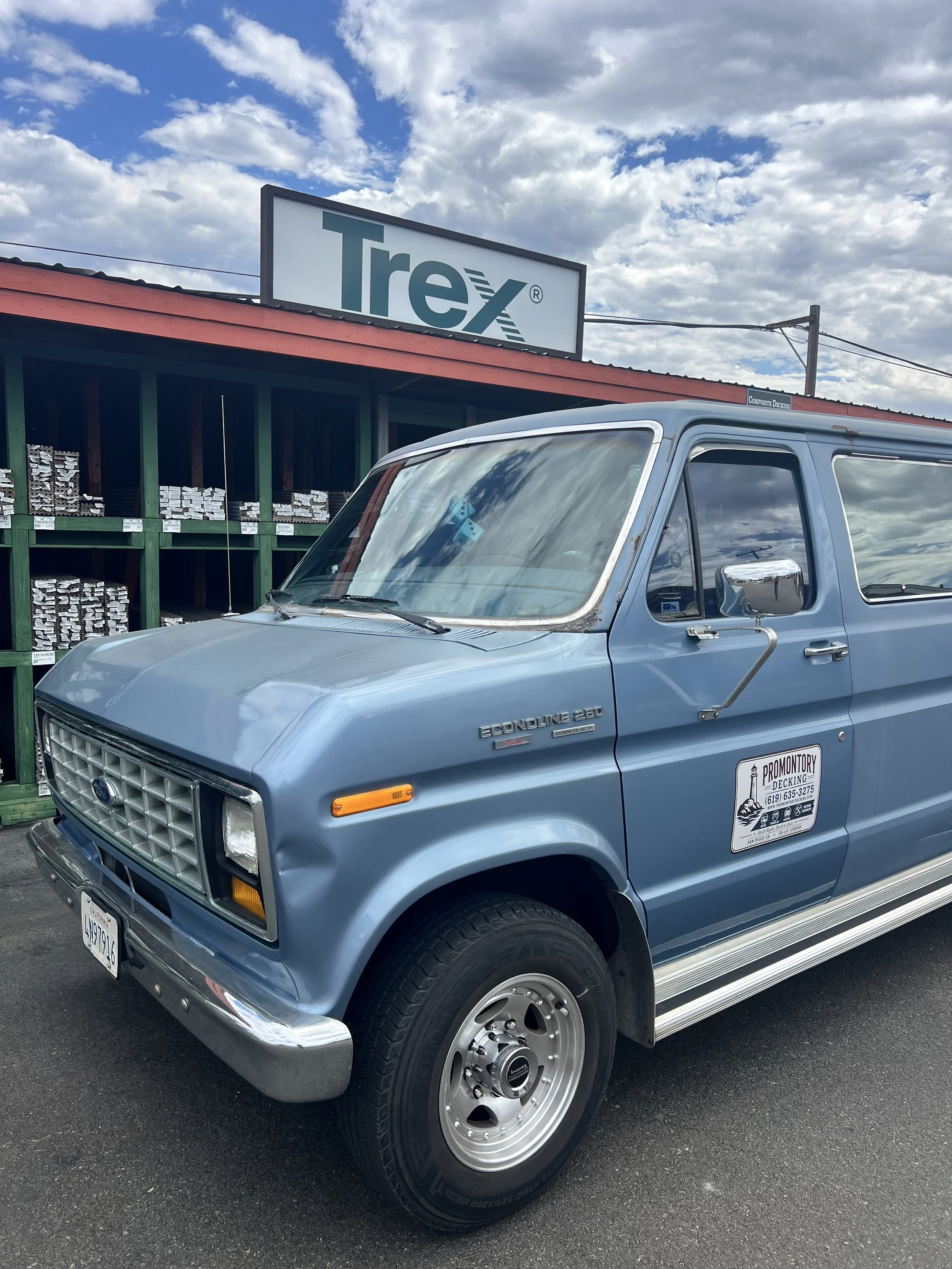 A vintage blue Ford Econoline van parked in front of a building with a sign that reads "Trefx", under a partly cloudy sky. The vehicle has a sticker on the side that reads “Promontory Decking,” and a California license plate.