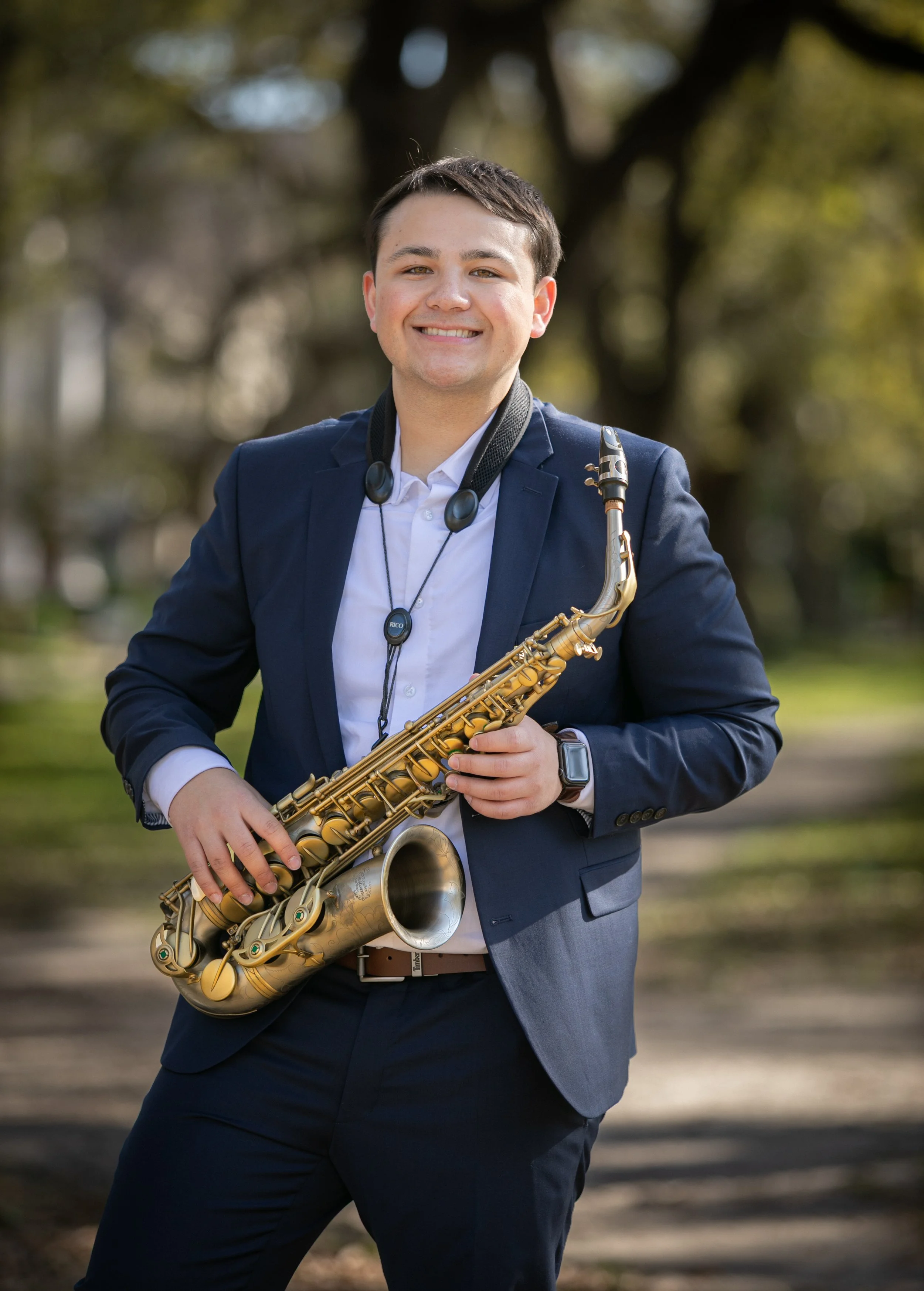 A young man in a navy blue suit holding a gold-colored saxophone outdoors, smiling at the camera with trees in the background.