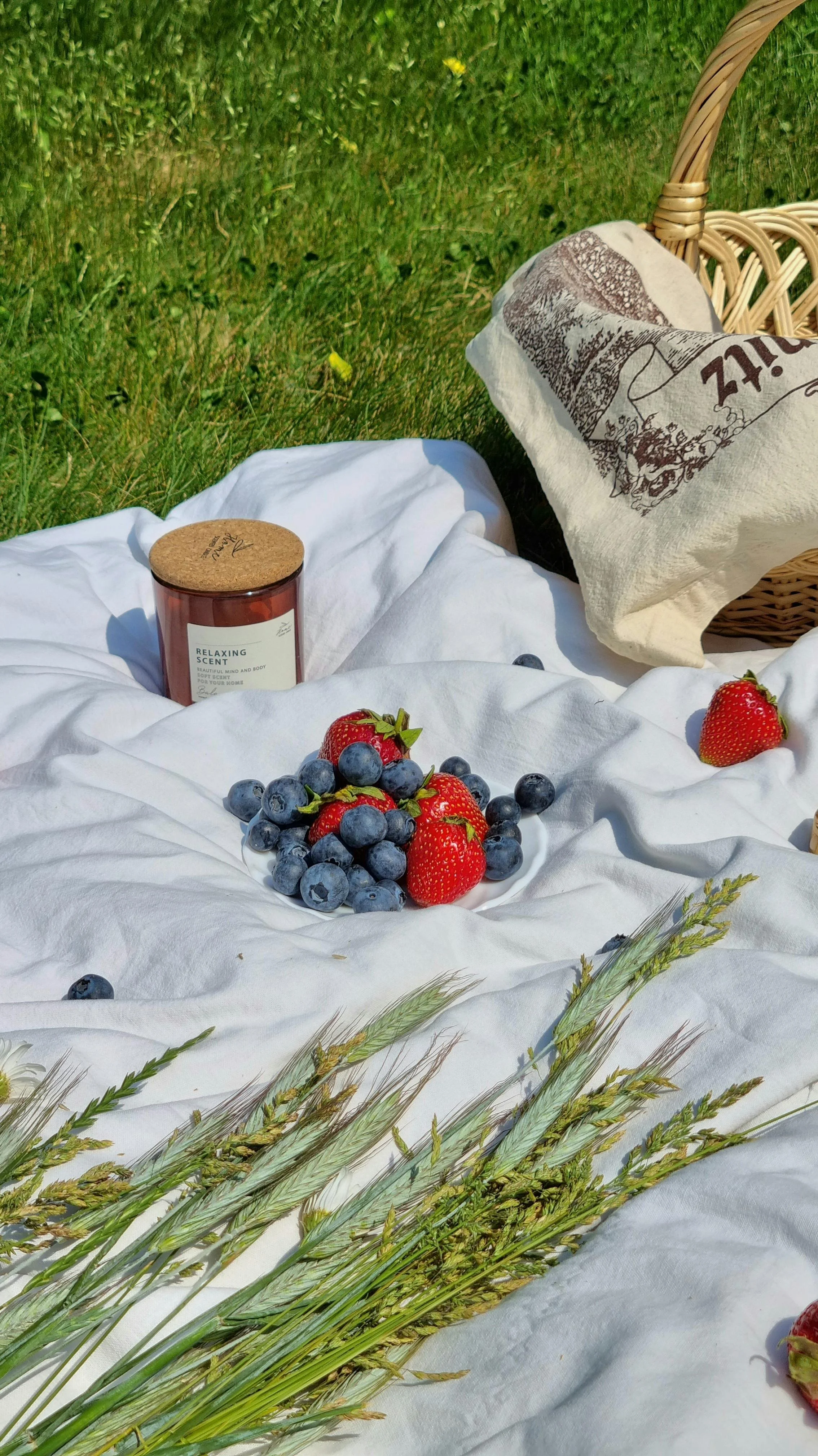 A picnic setup on white cloth with fresh strawberries, blueberries, a straw basket, a scented candle, and a bag with a printed design, on green grass.