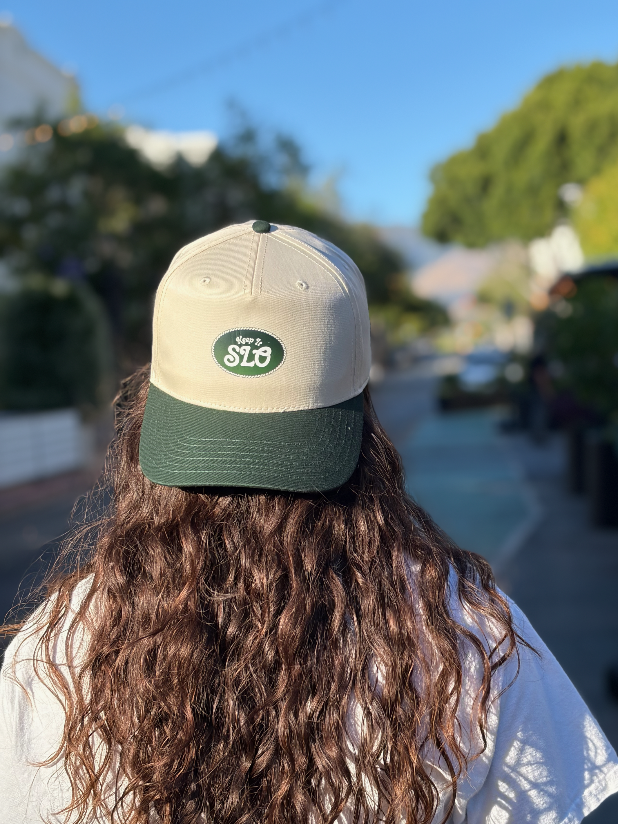 Person with long, curly brown hair wearing a beige and green baseball cap with the logo 'Keep It SLO' on the front, standing outdoors on a sunny day with trees and blue sky in the background.