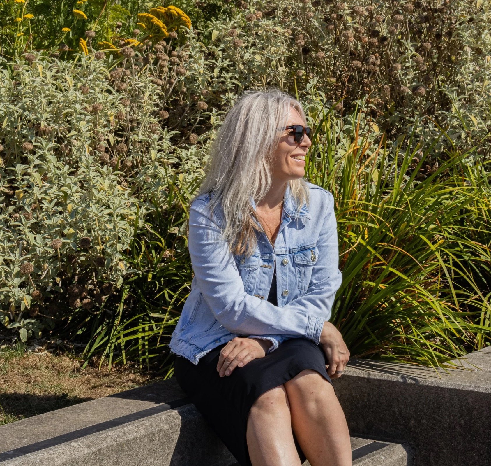 A woman with long gray hair wearing sunglasses, a light blue denim jacket, and a black dress, sitting on a stone bench outdoors surrounded by lush green plants and bushes, smiling and looking to her right.
