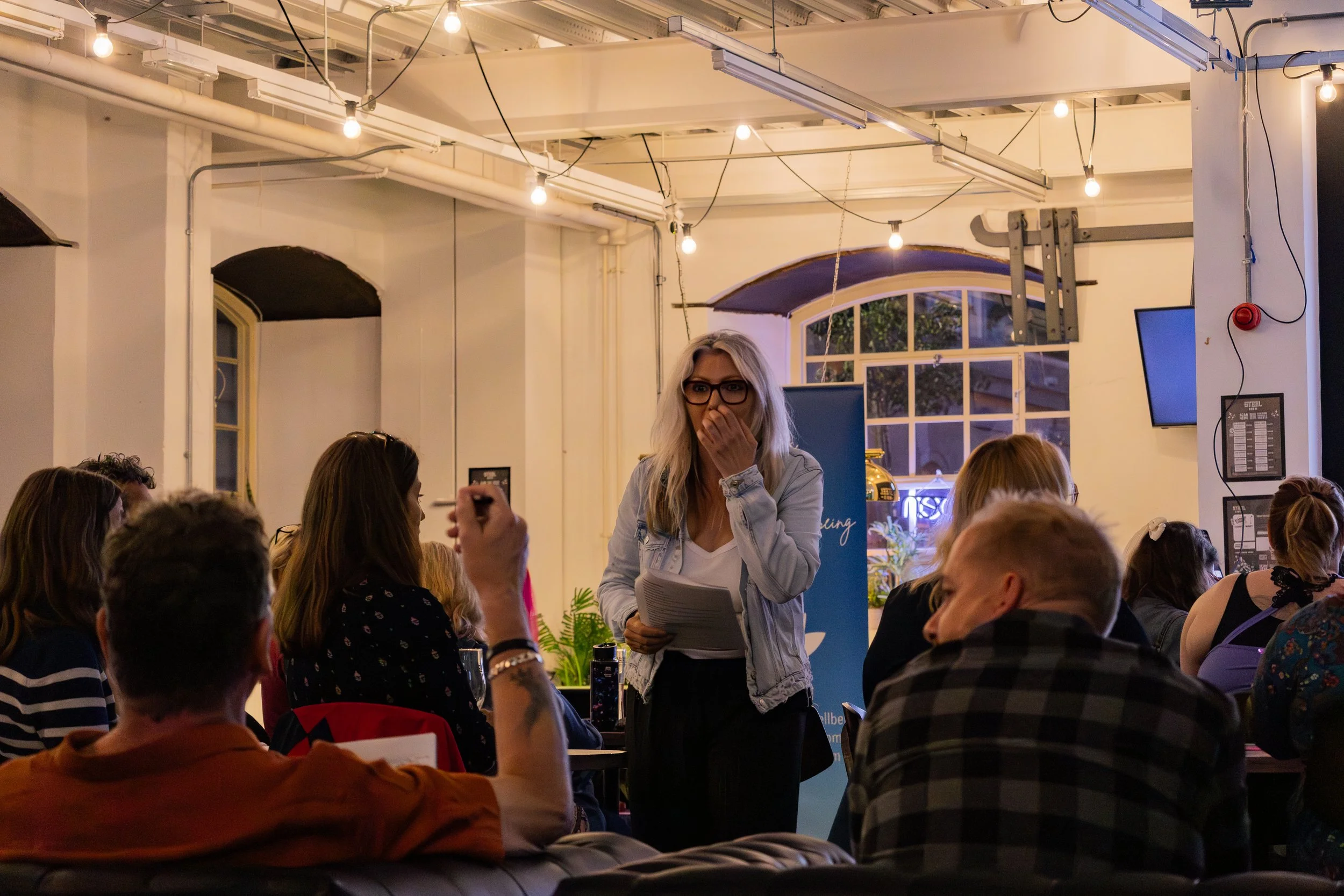 A woman with gray hair and glasses stands in front of a group of people in a cozy, well-lit room, holding papers and covering her mouth with her hand, appearing surprised or amused.