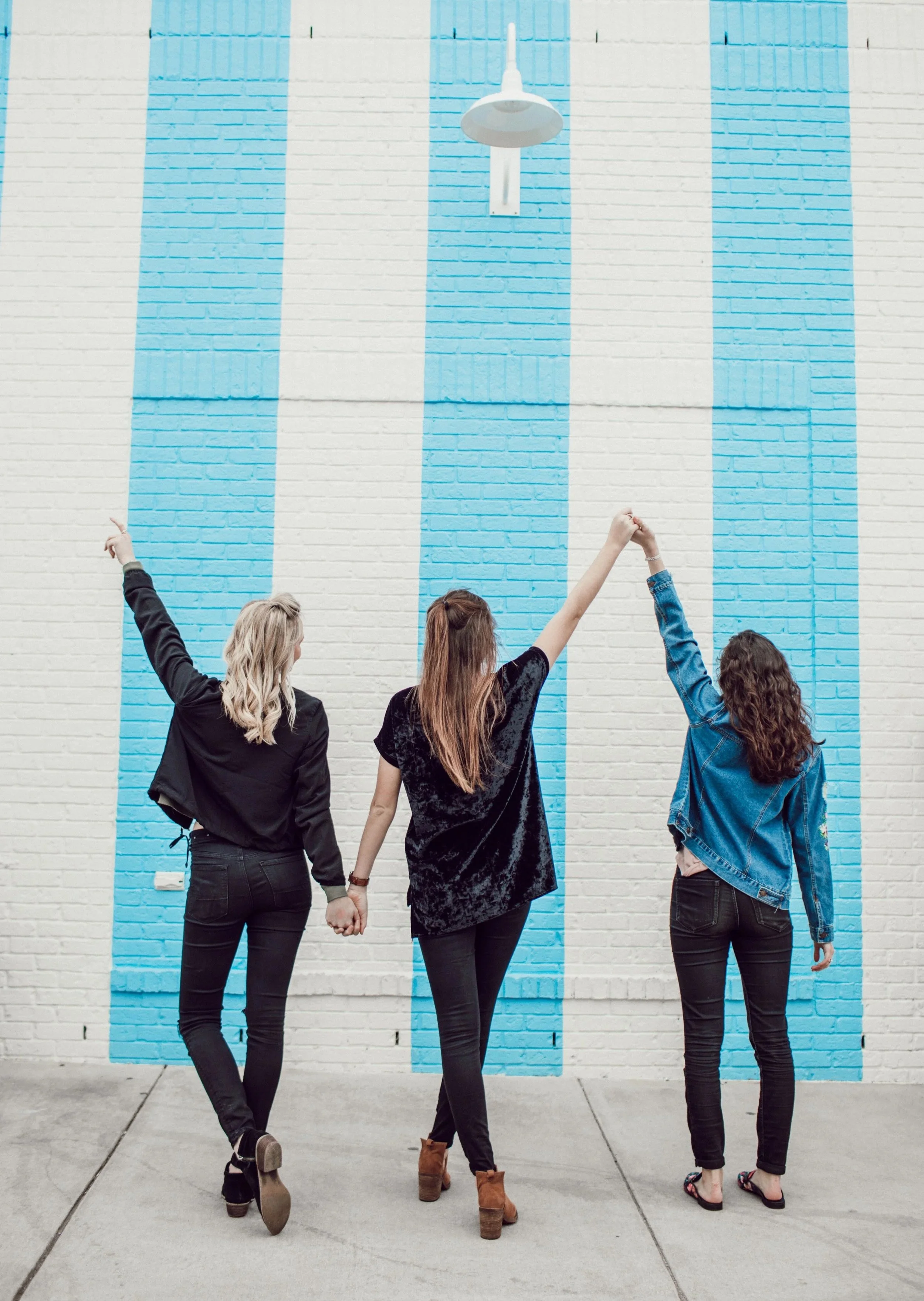 Three women hold hands and raise their arms, standing in front of a white brick wall with blue vertical stripes and a hanging light fixture.