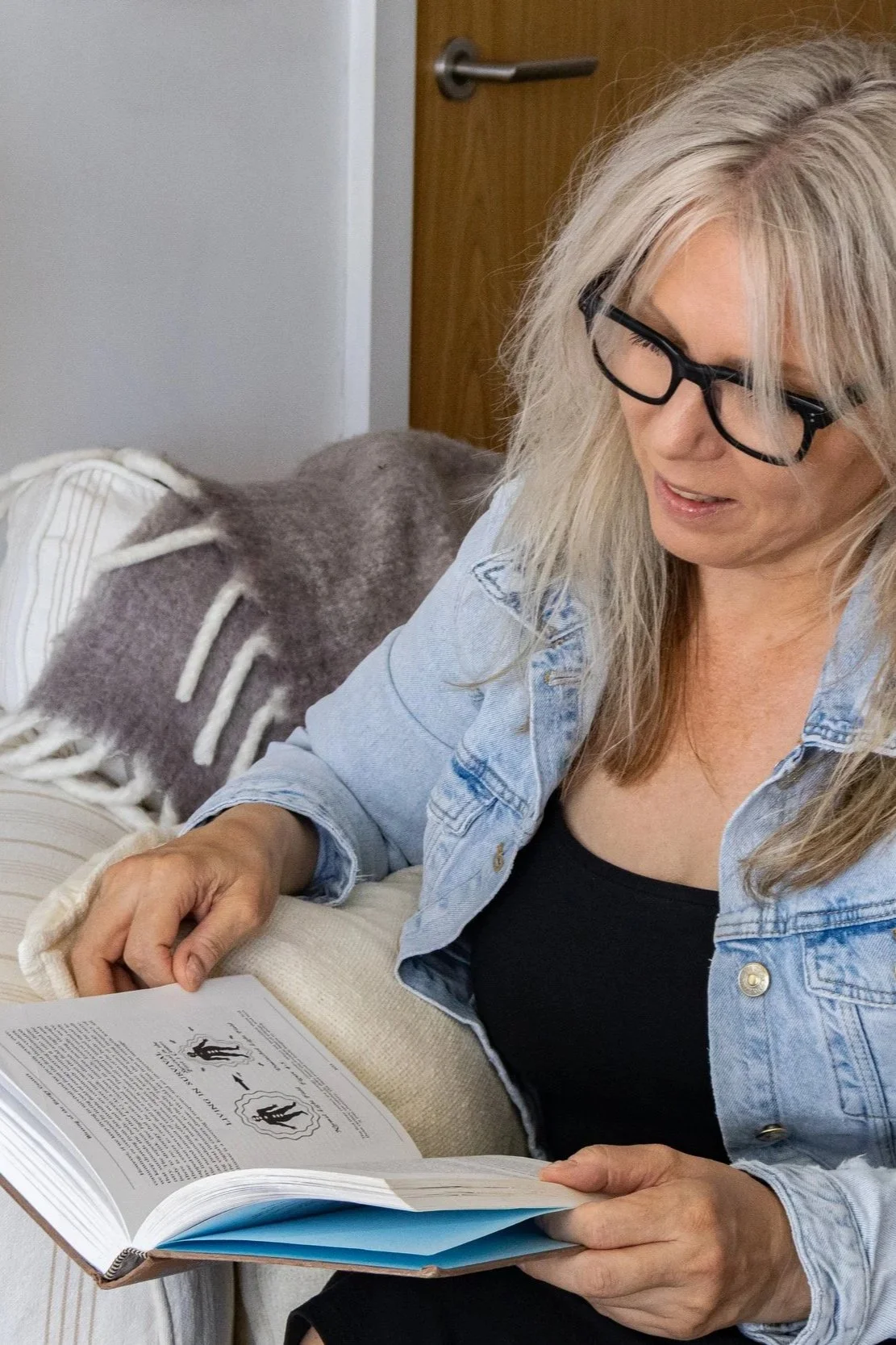 A woman with blonde hair, black glasses, and a denim jacket reading a book at home.