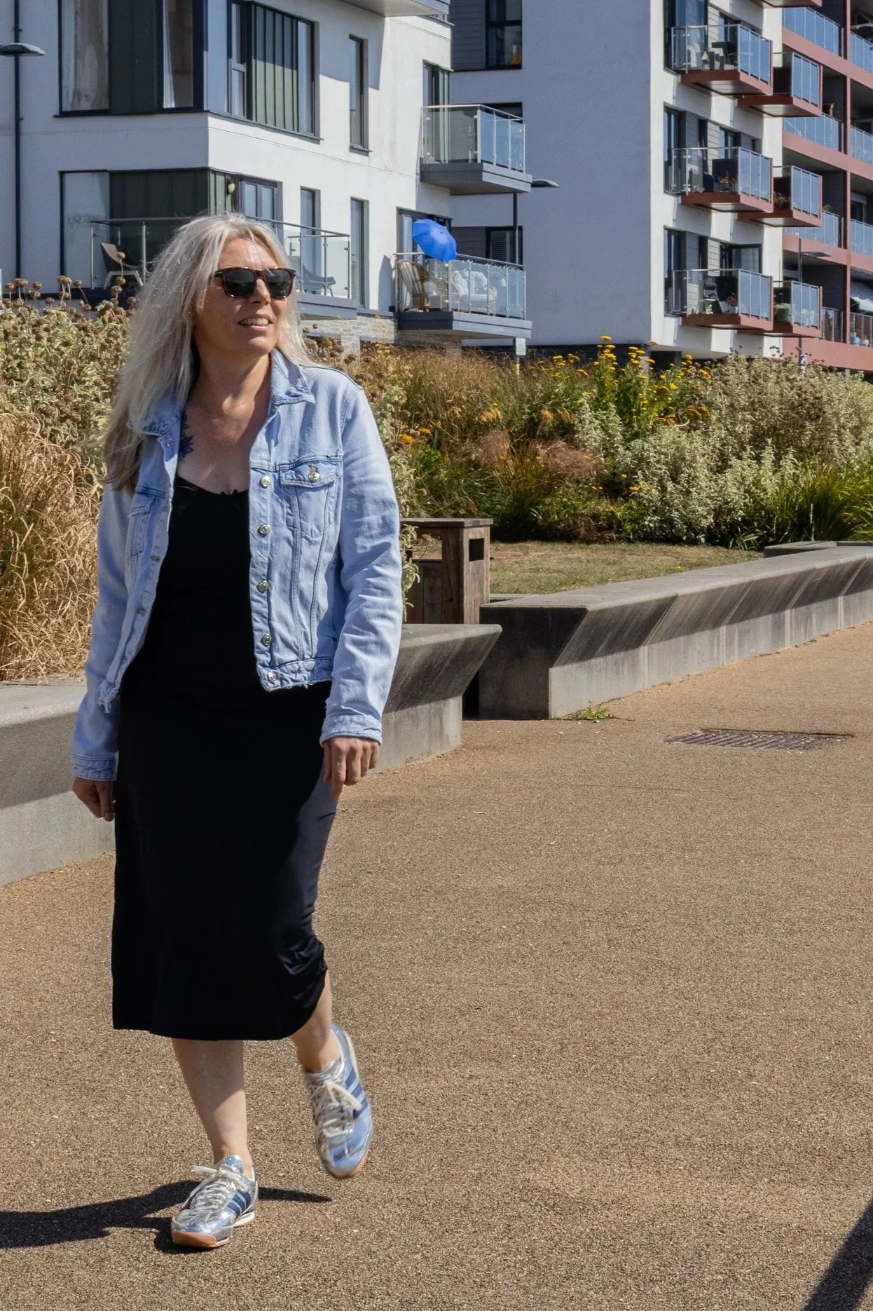 A woman with blonde hair wearing sunglasses, a light blue denim jacket, a black dress, and sneakers walks along a paved path near modern apartment buildings with balconies and a landscaped area with plants.