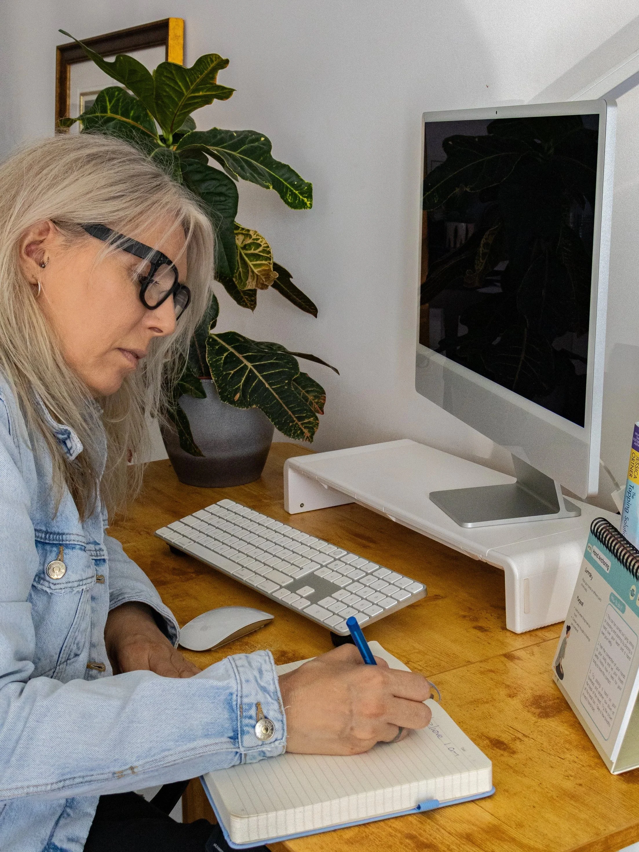 A woman with gray hair, glasses, and a denim jacket writing in a notebook at a wooden desk with a computer monitor, keyboard, and mouse, and a potted plant in the background.
