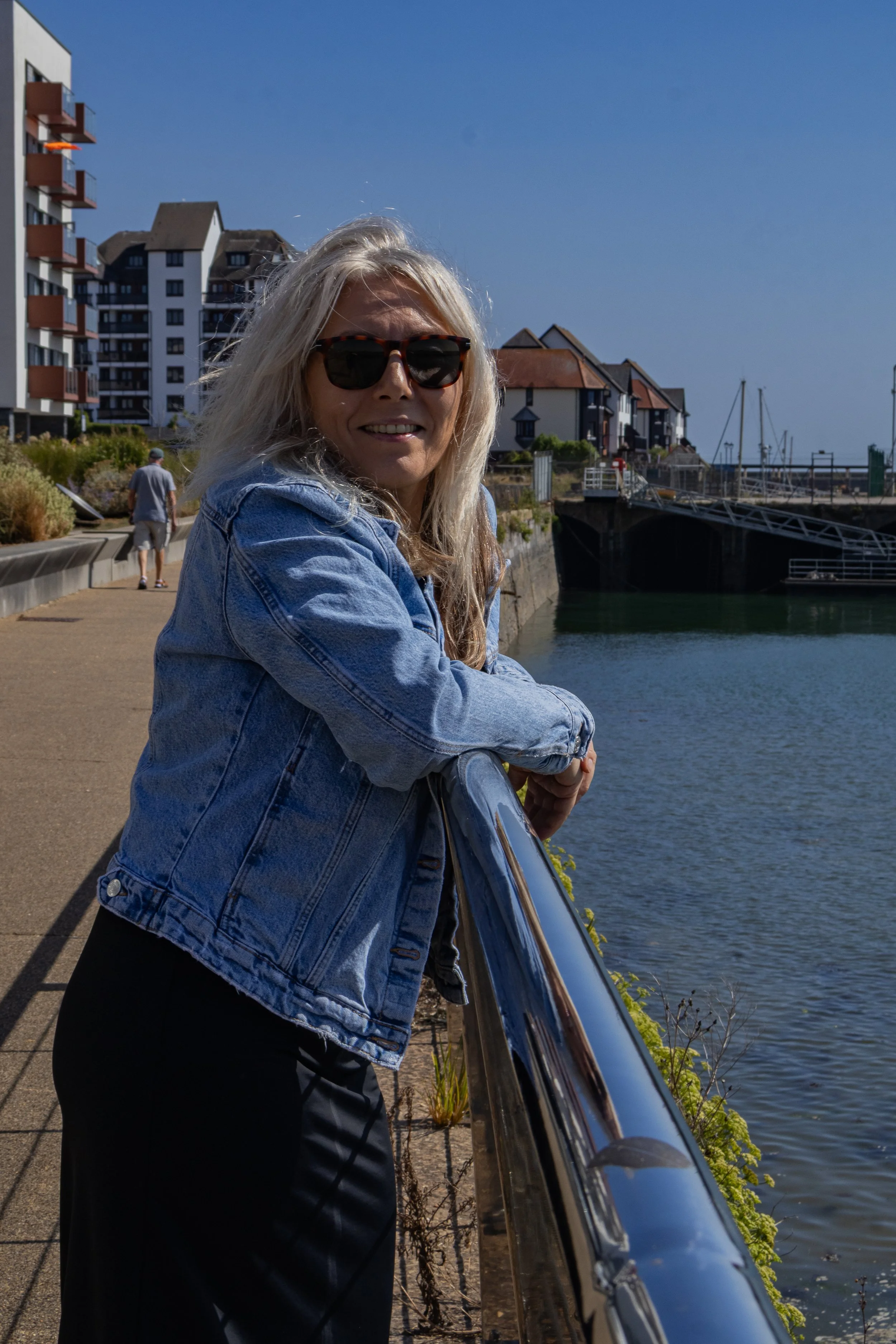 A woman with long blonde hair wearing a denim jacket and sunglasses leaning on a metal railing by the water, with buildings and boats in the background.