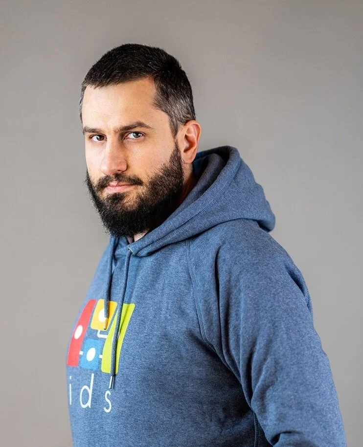 A man with short dark hair and a beard wearing a blue hoodie with colorful lettering standing against a plain gray background.