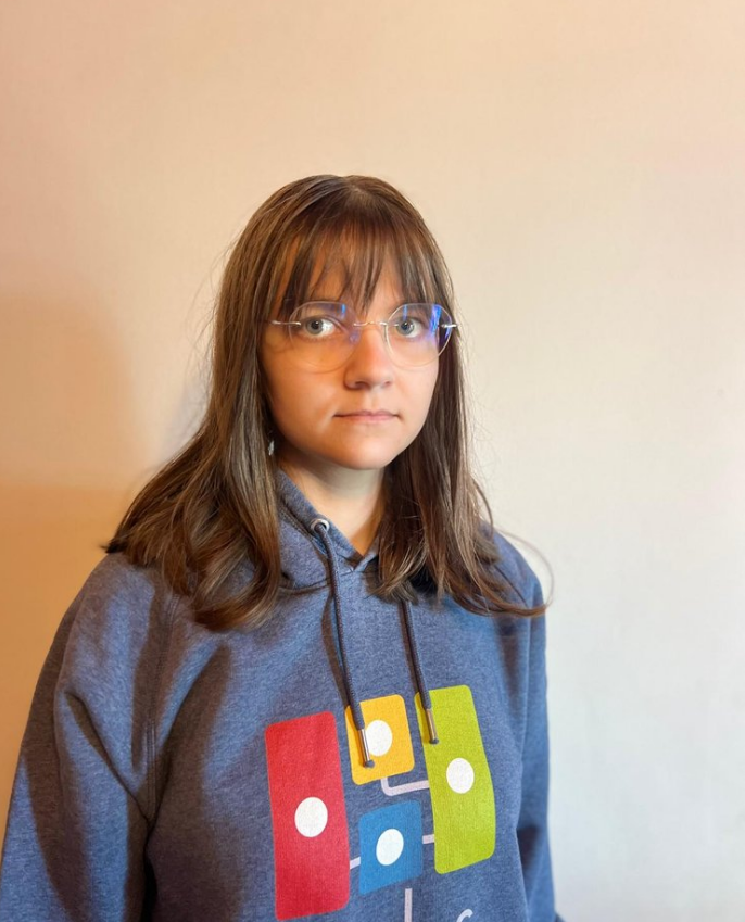 A young girl with brown hair, glasses, and a gray hoodie featuring colorful graphics, standing against a plain, light-colored wall.