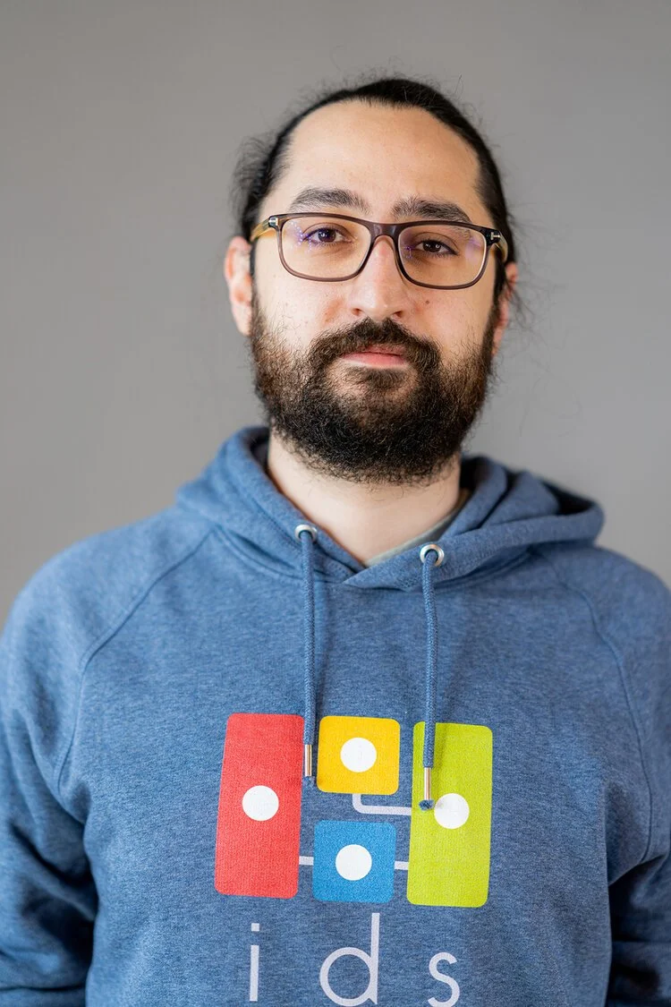 A man with glasses and a beard wearing a blue hoodie with a colorful dominoes graphic and the word 'ids' on it, standing against a gray background.