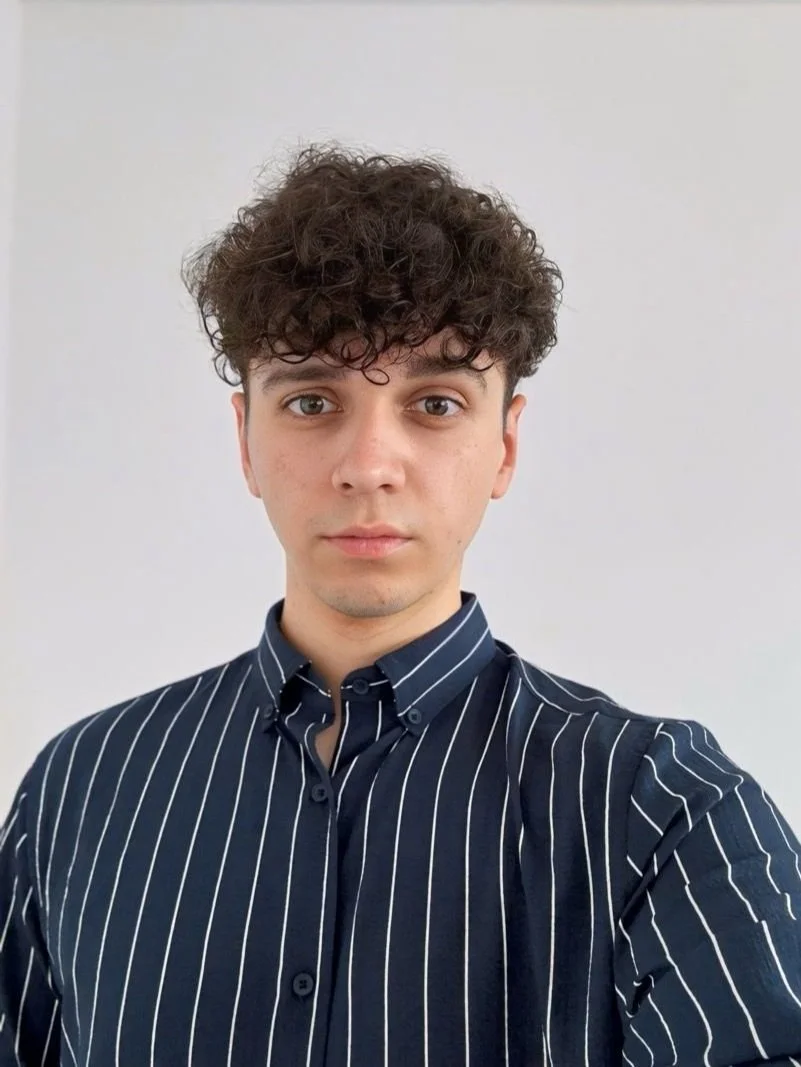 A young man with curly brown hair wearing a black shirt with white vertical stripes, taking a selfie in a plain white room.