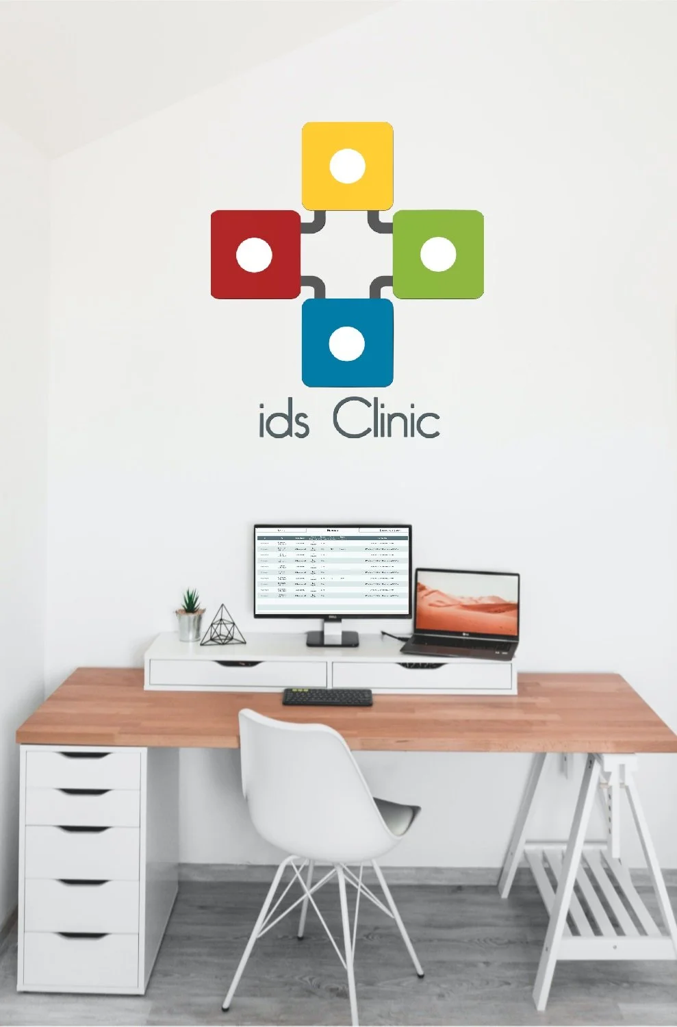 Office workspace with a wooden desk, a computer monitor, a laptop, and decorative items, against a white wall with a logo that says 'ids Clinic' and a colorful design of four connected squares.