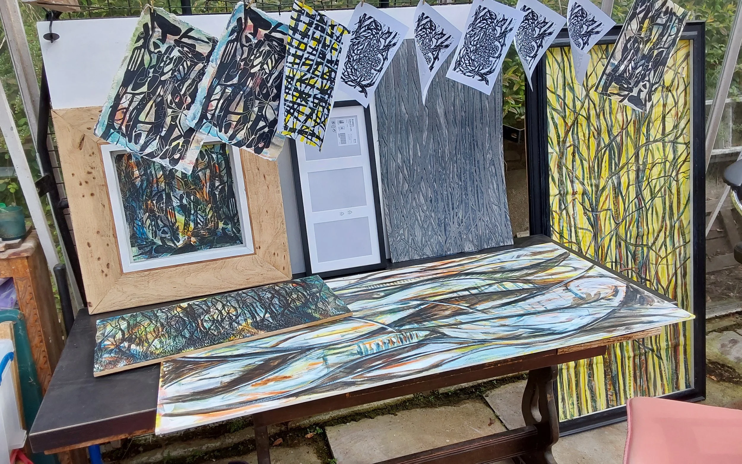 Outdoor display of various abstract and nature-inspired paintings, sketches, and framed artworks arranged on a black table with some hanging on a string of paper art.