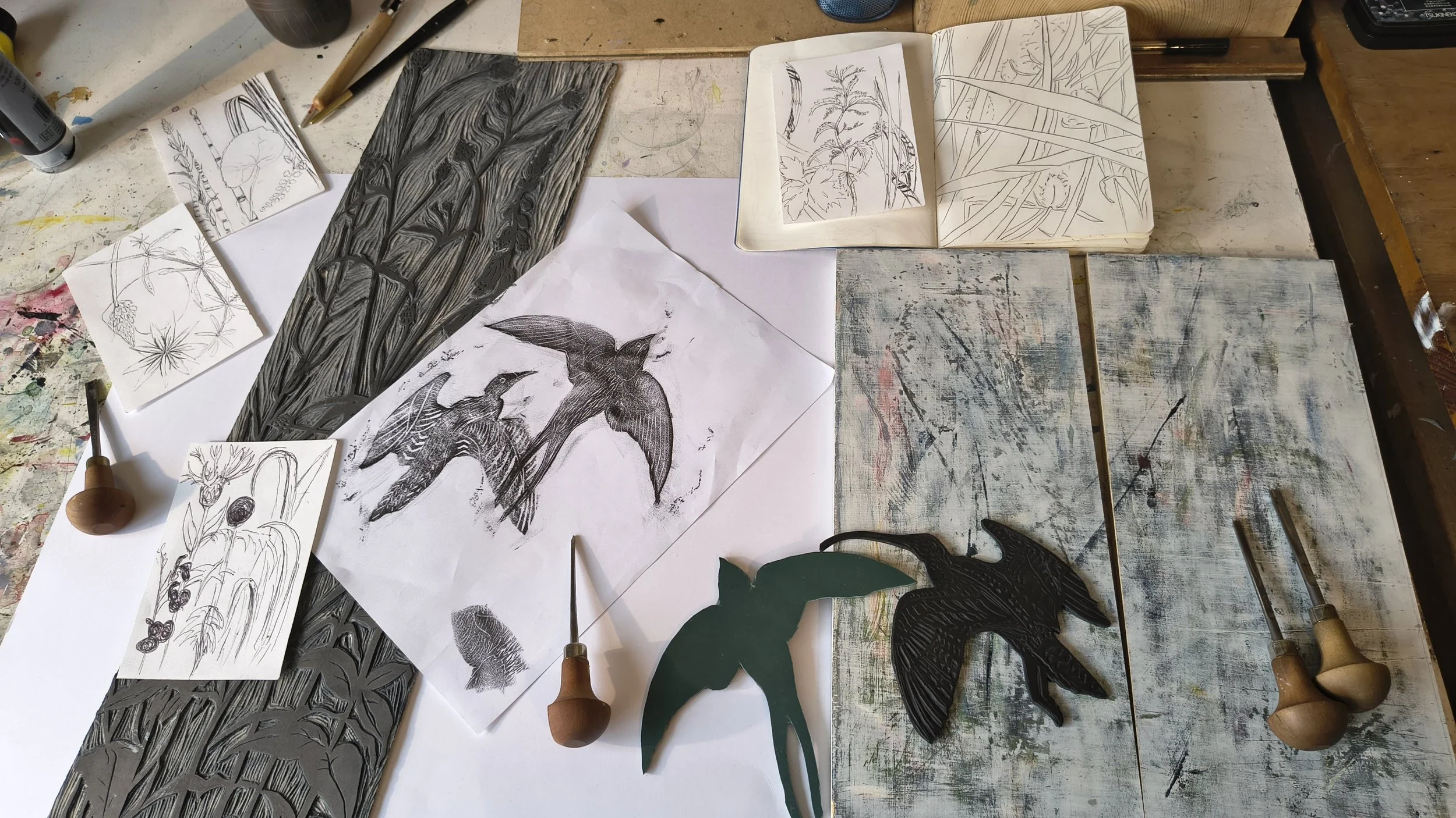 Workspace with sketches, carvings, and tools of birds and plant designs for printmaking or woodworking projects. Includes sketches, carved wood blocks, and carving tools.