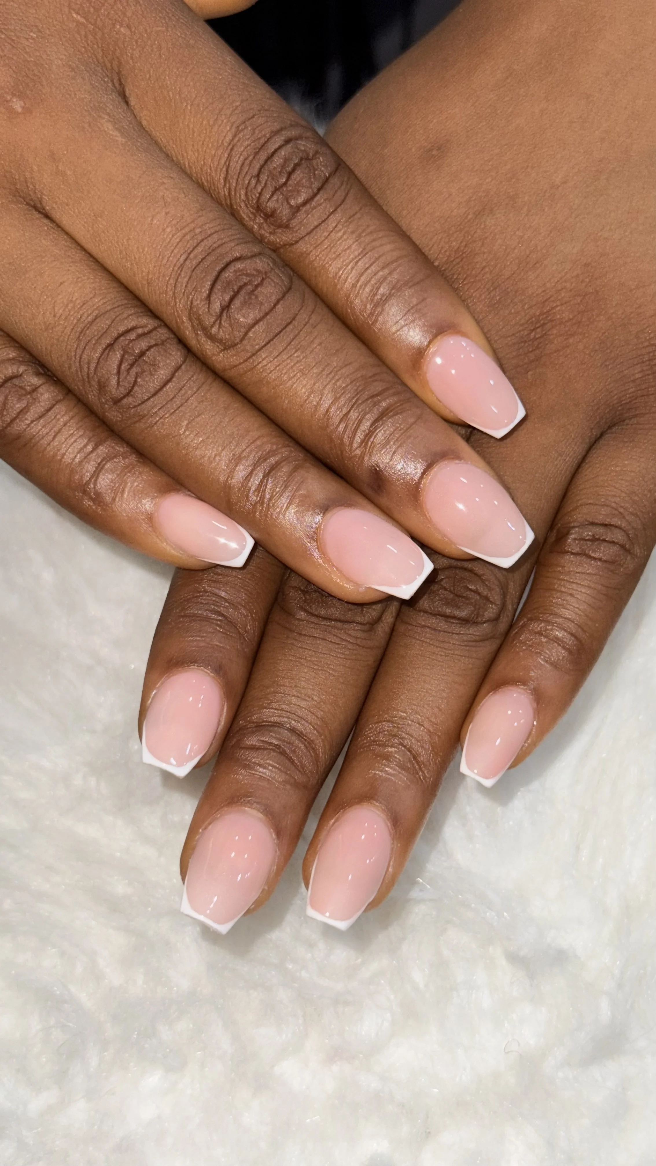 Close-up of hands with manicured nails, featuring natural skin tone. Nails are shaped in a square style with a French manicure, light pink base, and white tips. The background has a white textured surface.