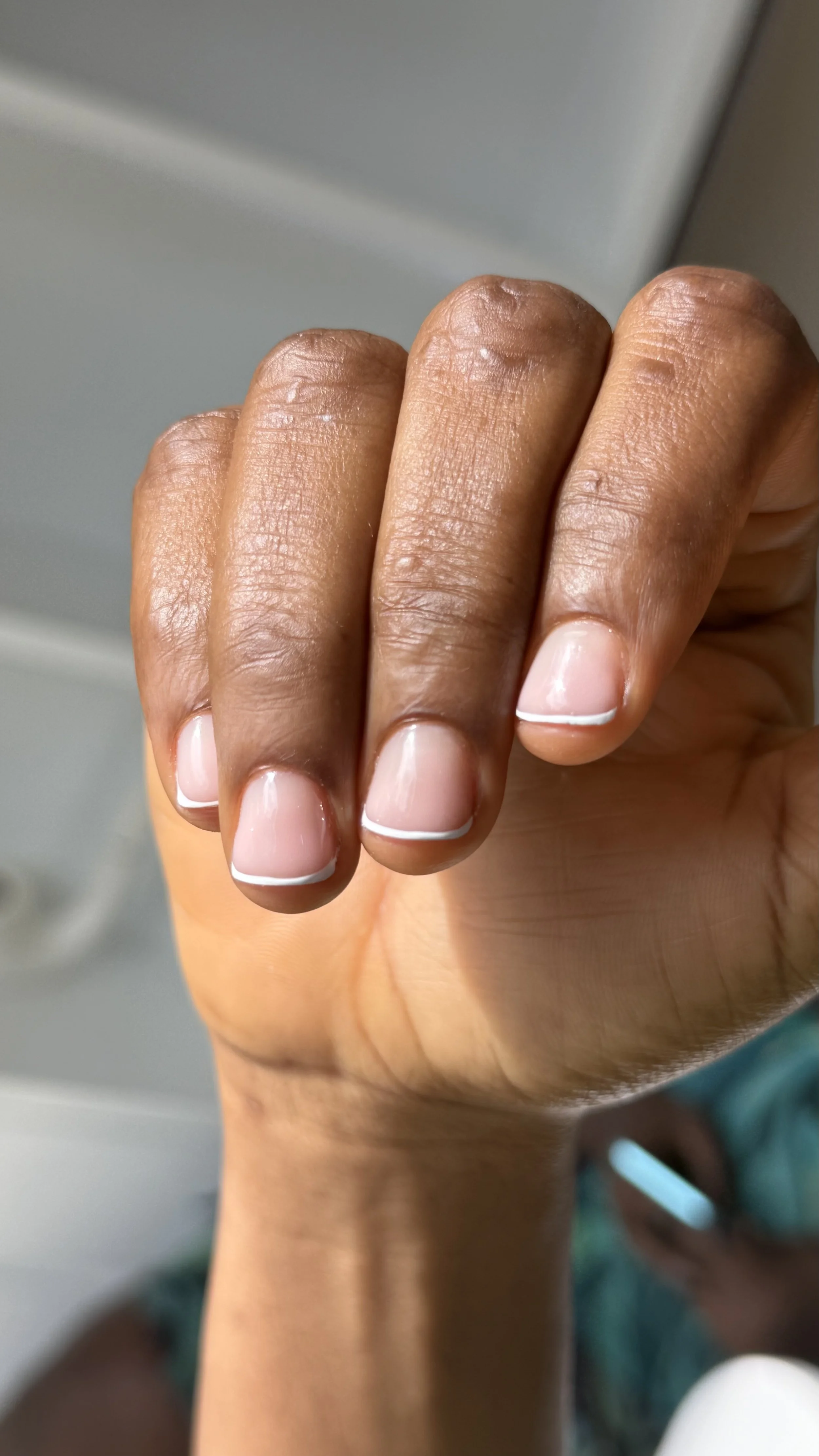 Close-up of a person's hand with a darker skin tone, showing well-manicured nails with a natural look and a white tip, against a blurred background.