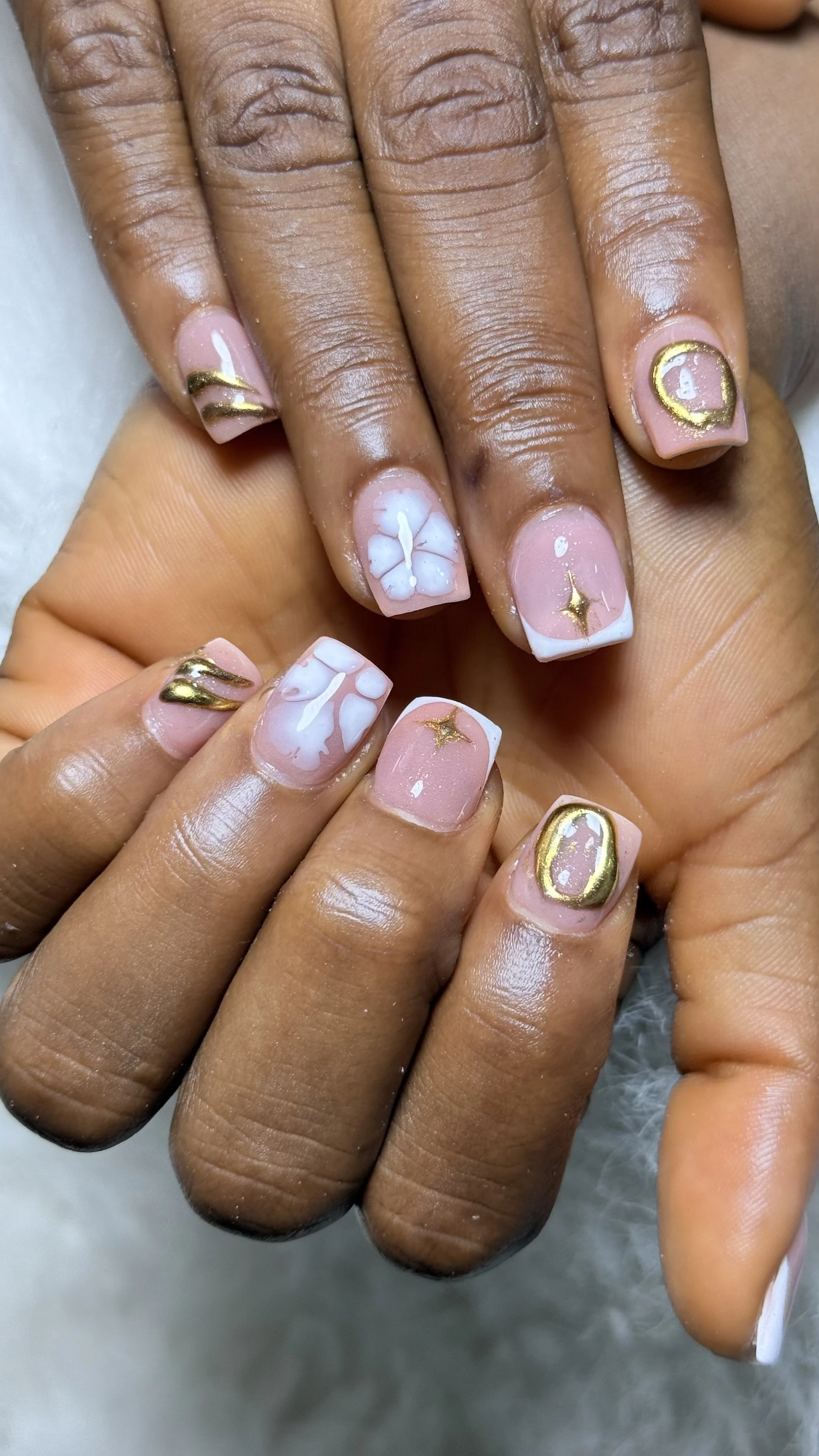 Close-up of a person's hands with manicured nails featuring a pink and white floral design, gold accents, and decorative patterns.