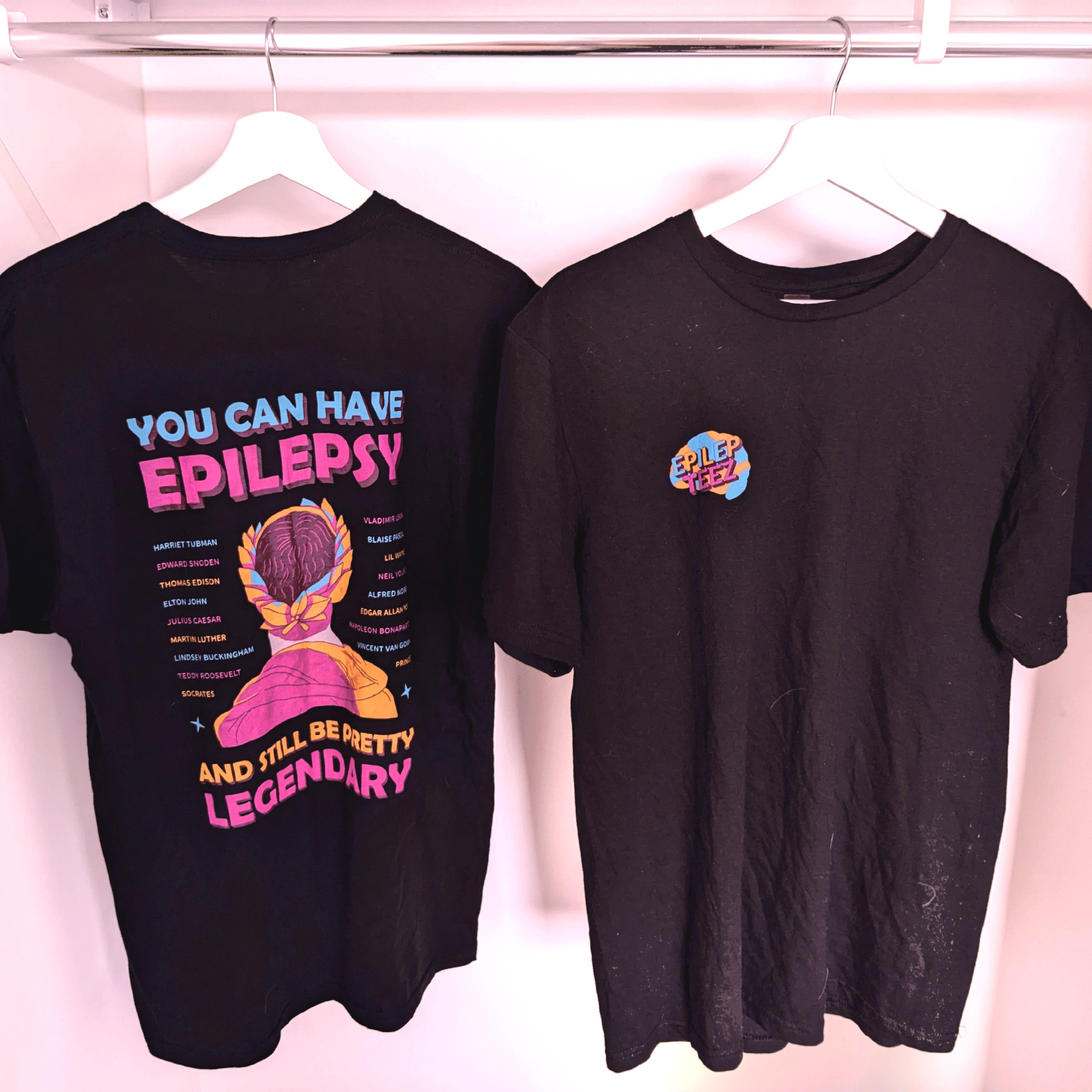 Two black T-shirts hanging on white hangers inside a closet, one with colorful text and graphics about epilepsy and the other with a small colorful patch that says "EPILEPTEEZ."