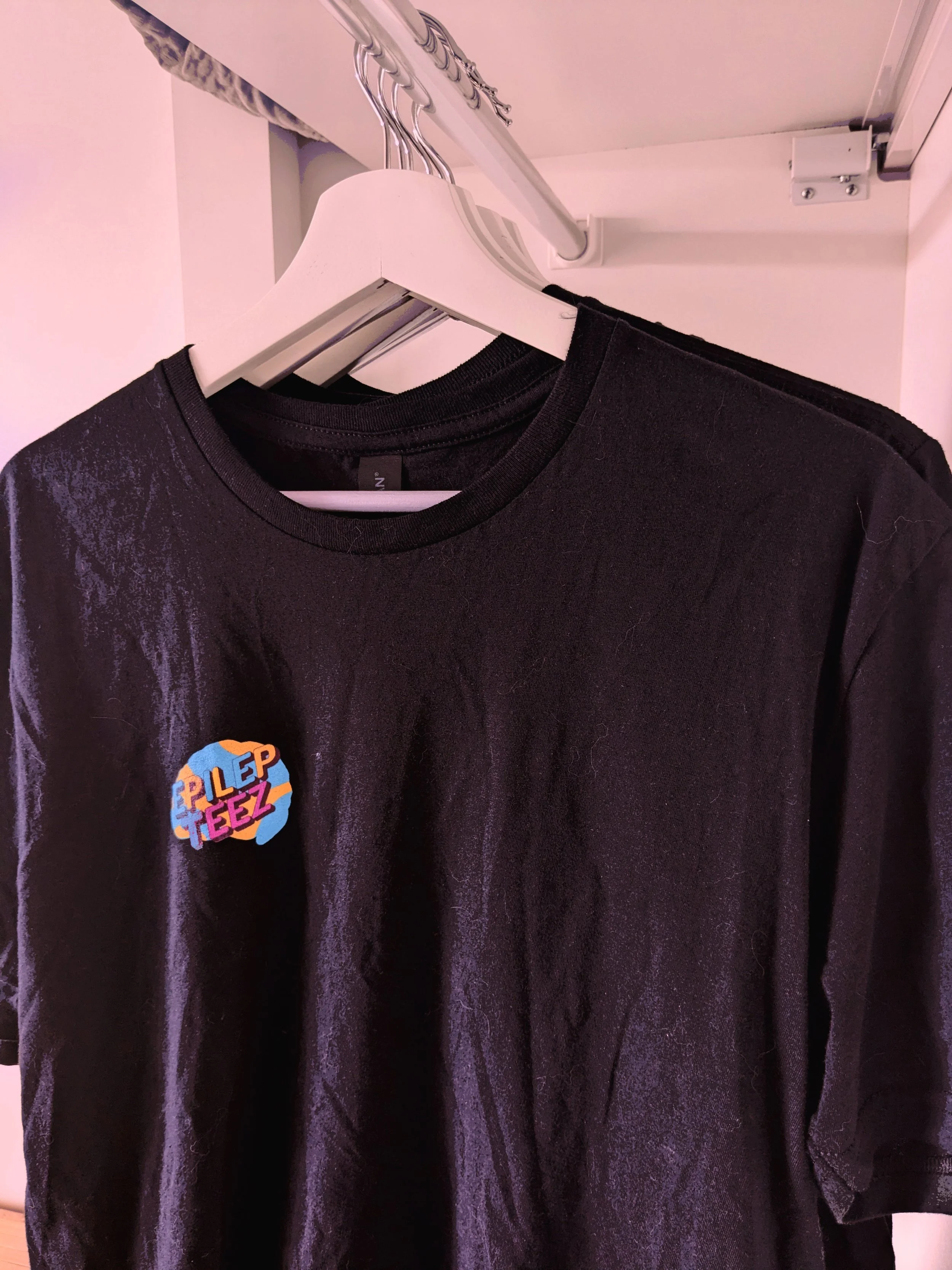 Black t-shirt hanging on a white hanger inside a closet, featuring a colorful sticker that says 'PLEDGE' with vibrant, playful font and background.
