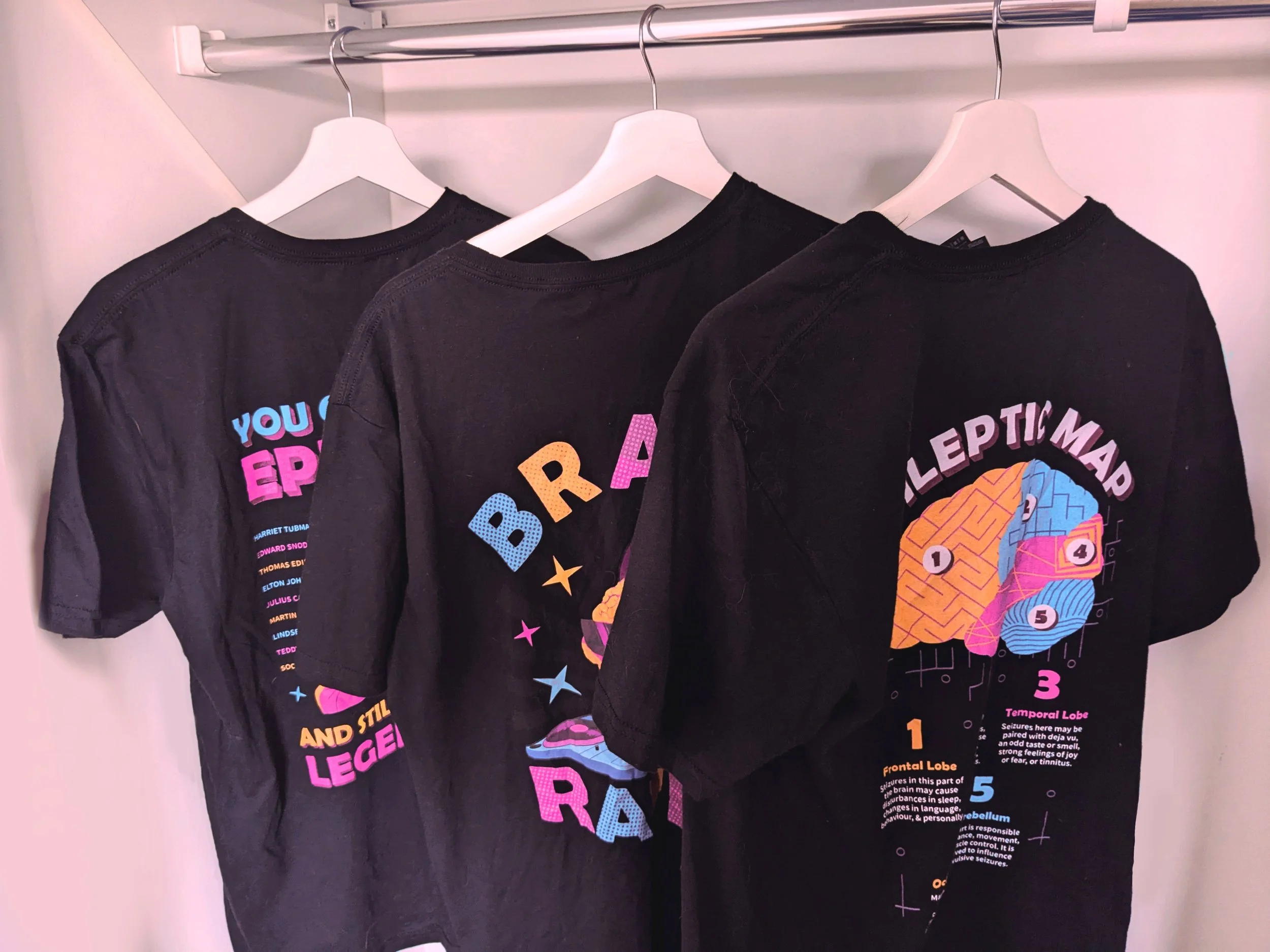 Three black T-shirts with colorful graphic designs and text about the brain, hanging on white hangers in a closet.