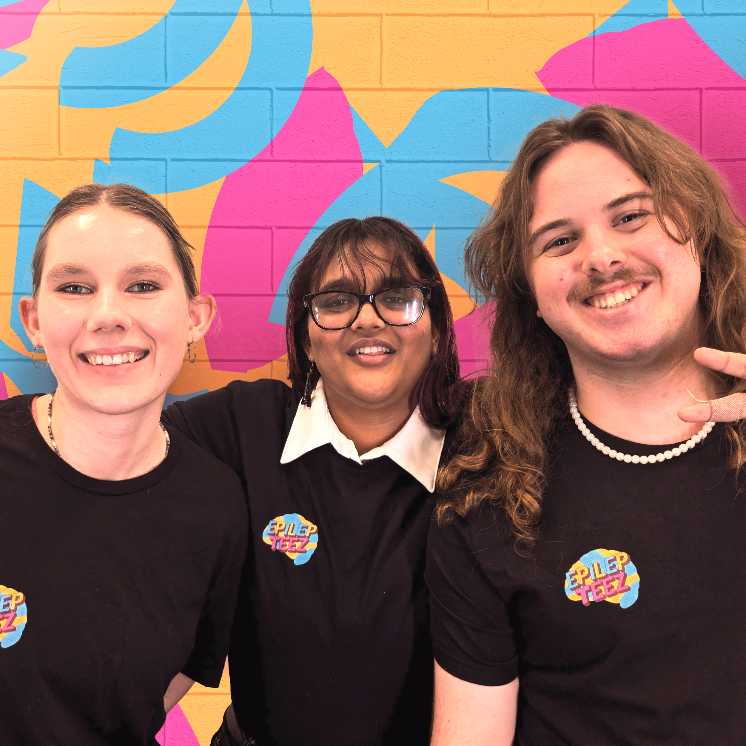 Three smiling people standing in front of a colorful graffiti wall wearing black t-shirts with a logo.
