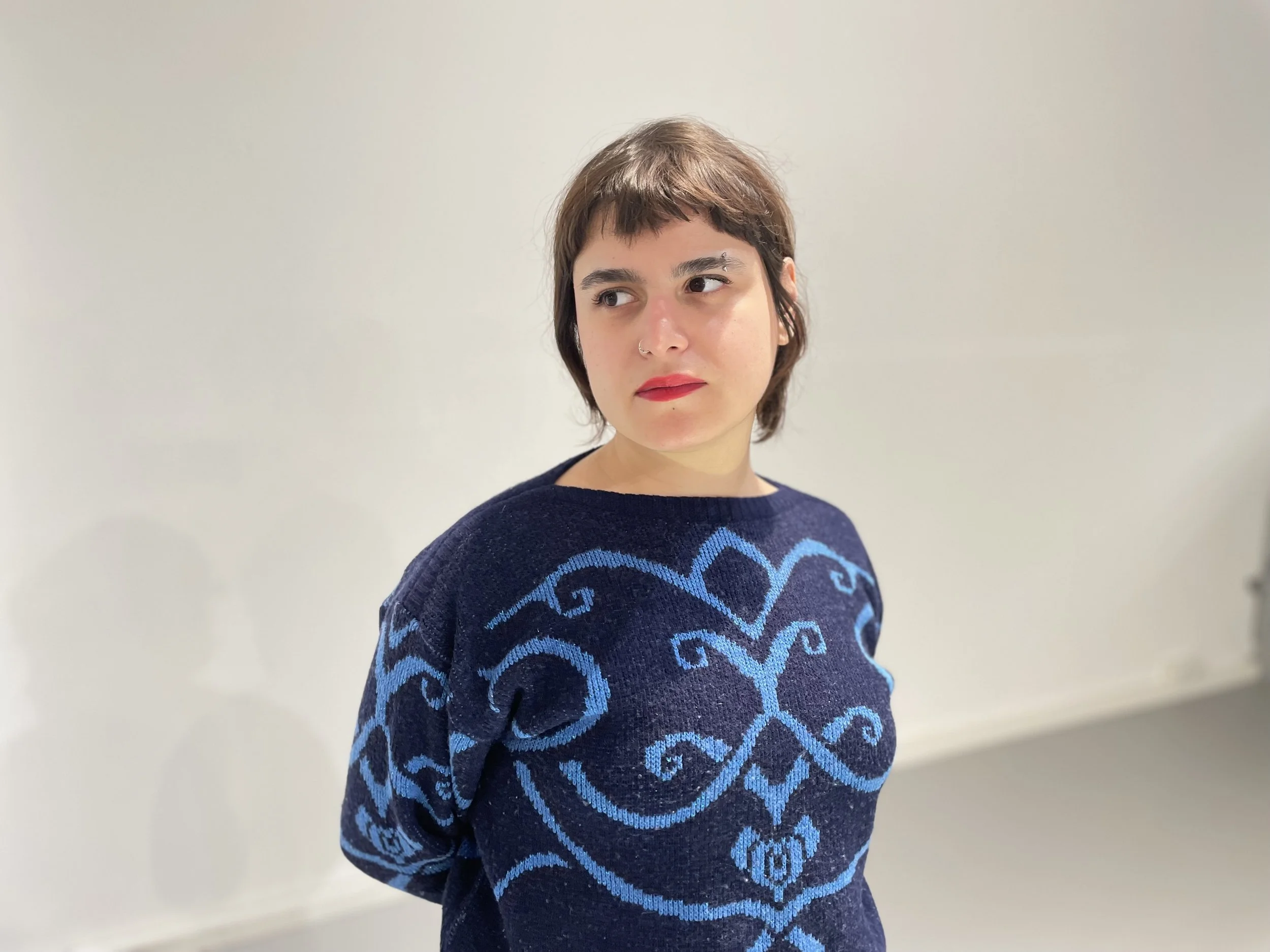 Young woman with short brown hair wearing a navy sweater with blue ornate patterns, standing against a plain off-white wall.