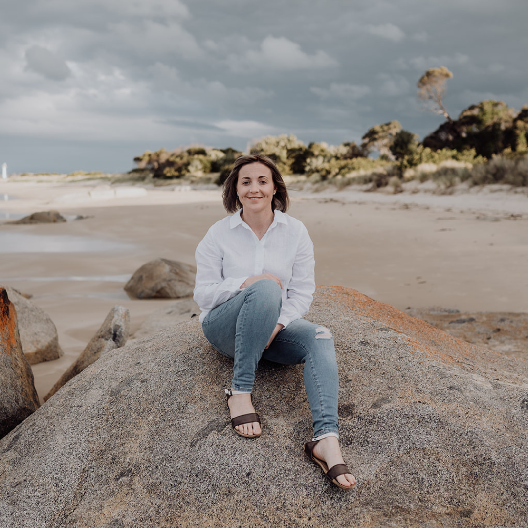 A woman wearing a white shirt and ripped jeans sitting on a large rock on a sandy beach with trees and cloudy sky in the background.