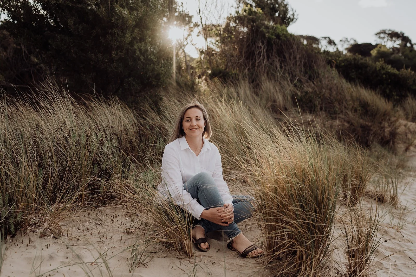 A woman sitting on the sand between tall beach grasses with trees and the sun in the background, smiling and wearing a white shirt and jeans.