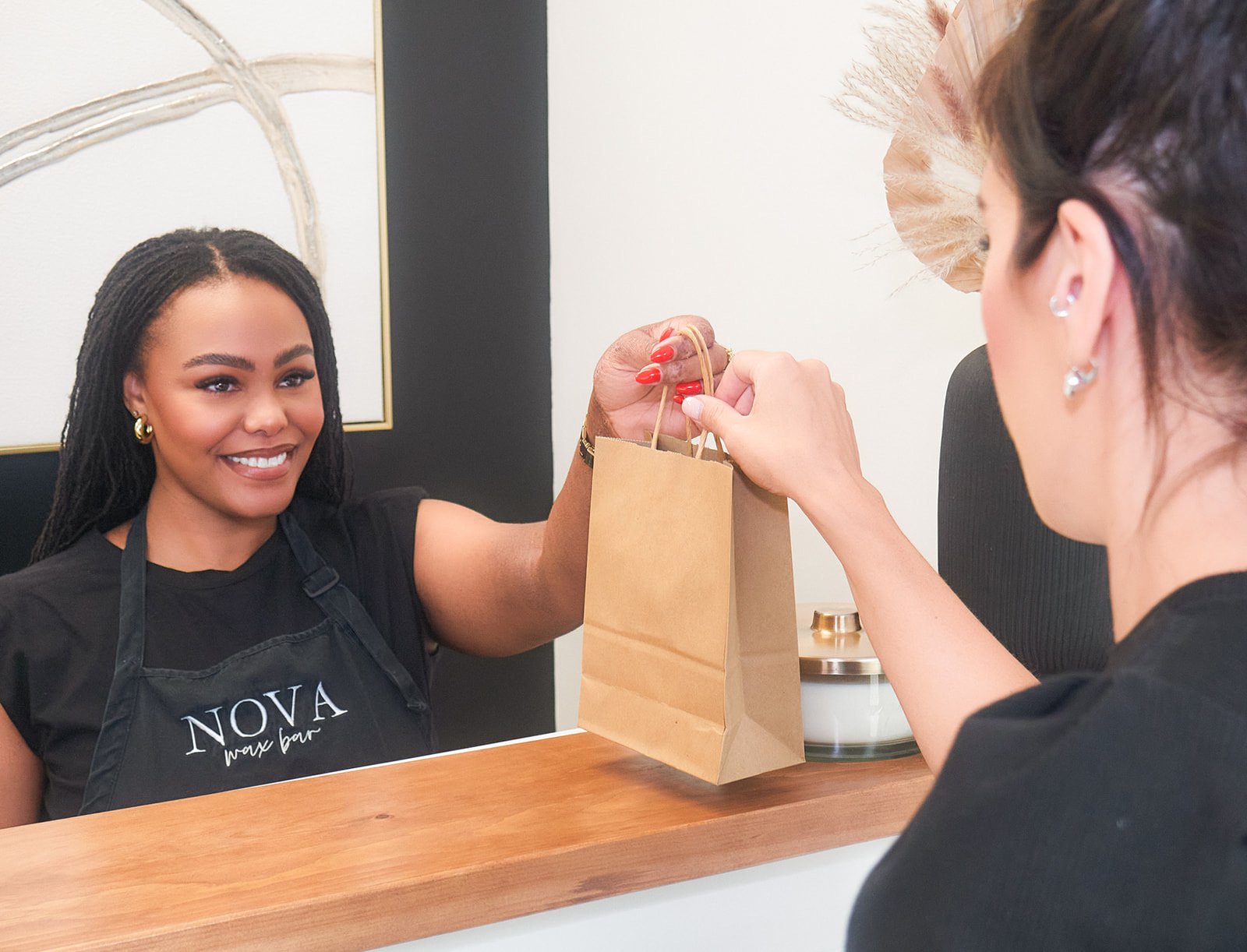 A woman with dark hair and teardrop earrings is handing over a paper bag to another woman at a counter. The woman behind the counter is smiling and wearing a black apron with white text that says "NOVA wax bar."