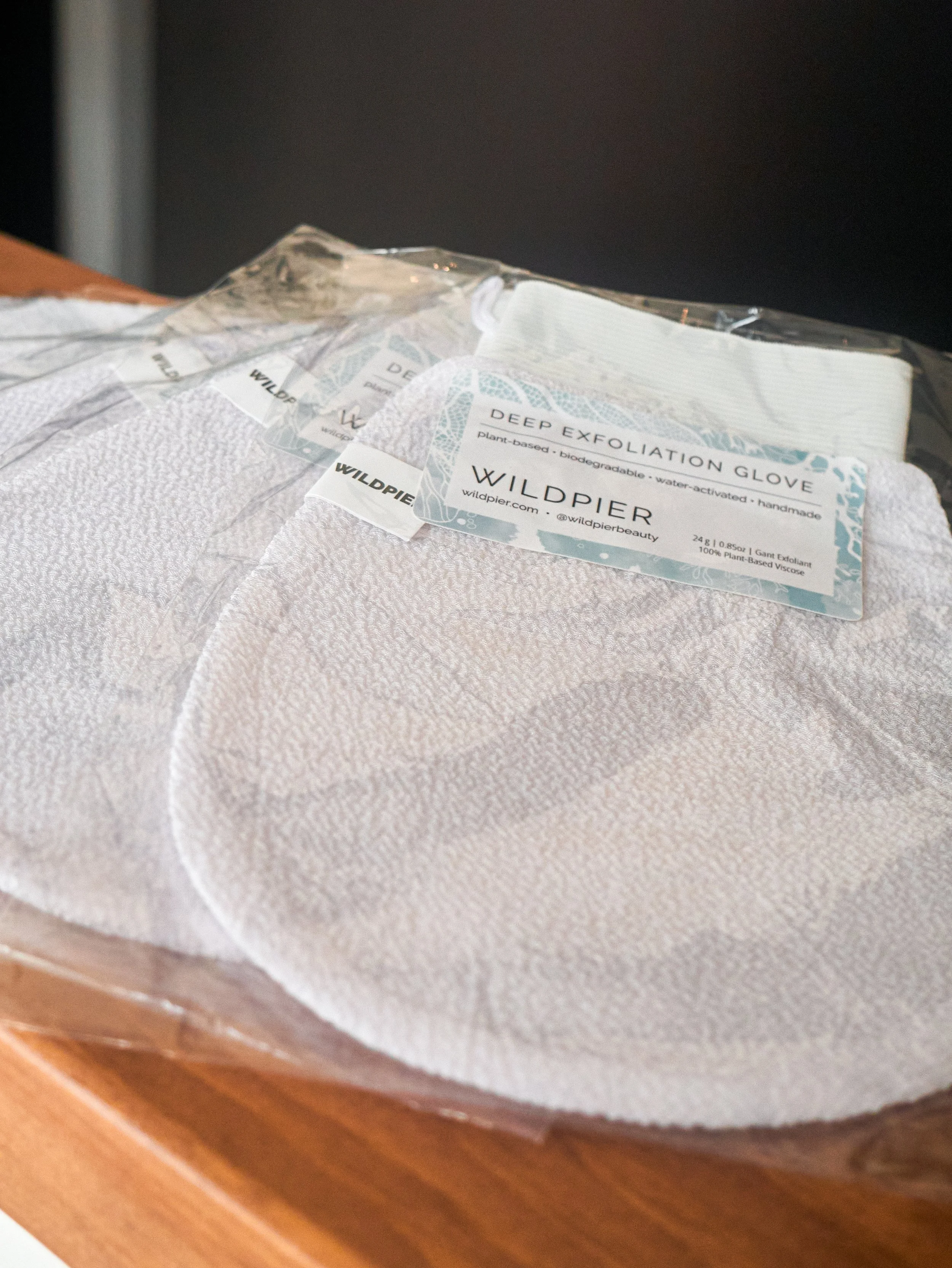 White exfoliation gloves in plastic packaging with labels from Wildpier, on a wooden surface.