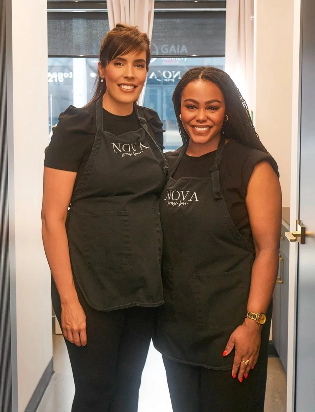 Two women in black aprons standing inside a restaurant kitchen, smiling at the camera.