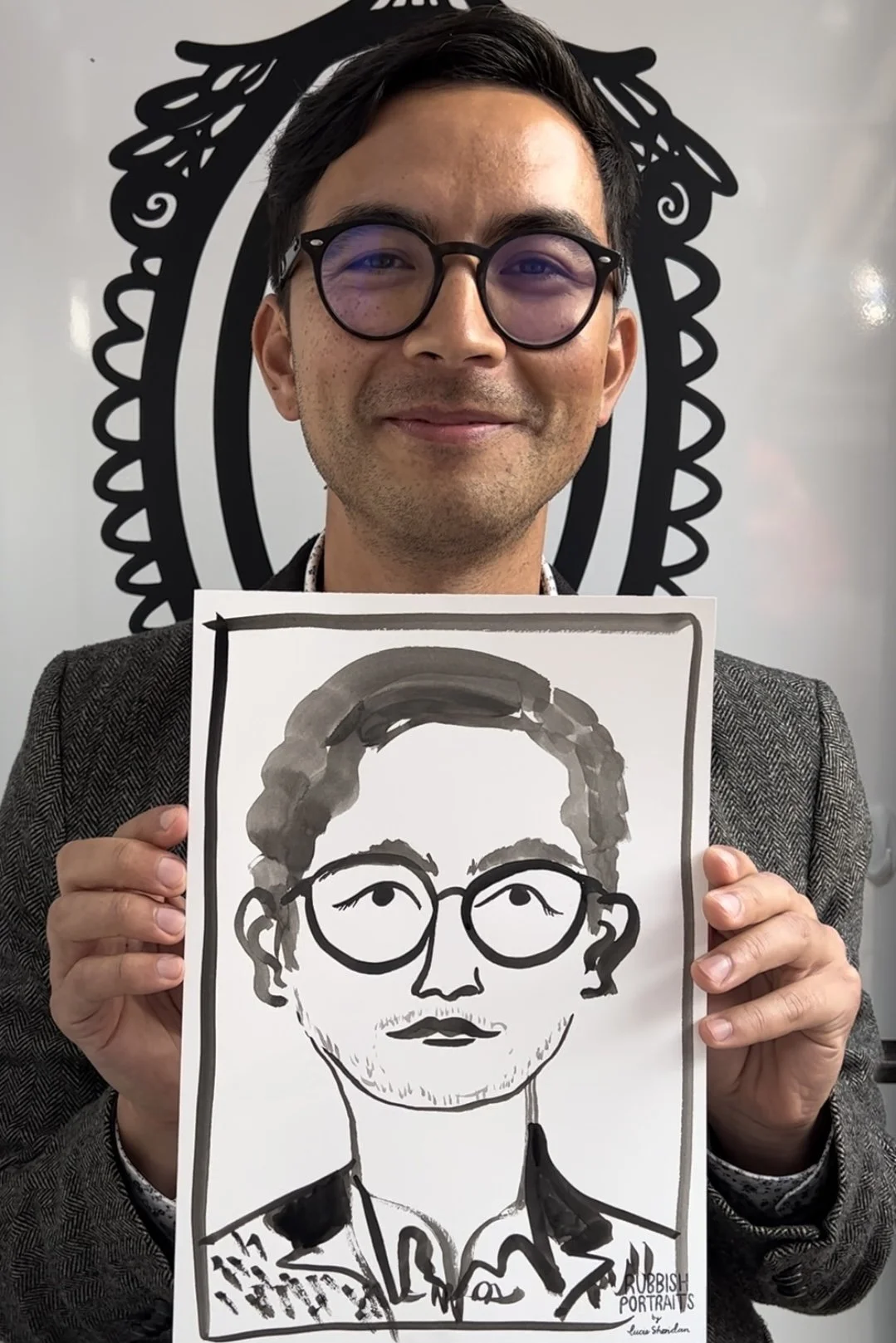 A man with black hair, glasses, and a gray blazer is holding a black-and-white Rubbish Portrait drawing of himself. The man is smiling and standing in front of a background with a decorative black and white design.