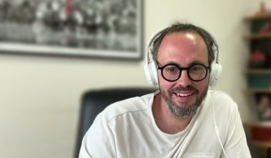 Homme souriant avec des lunettes et des écouteurs blancs, assis à un bureau dans une pièce avec un tableau ou une photo en arrière-plan.