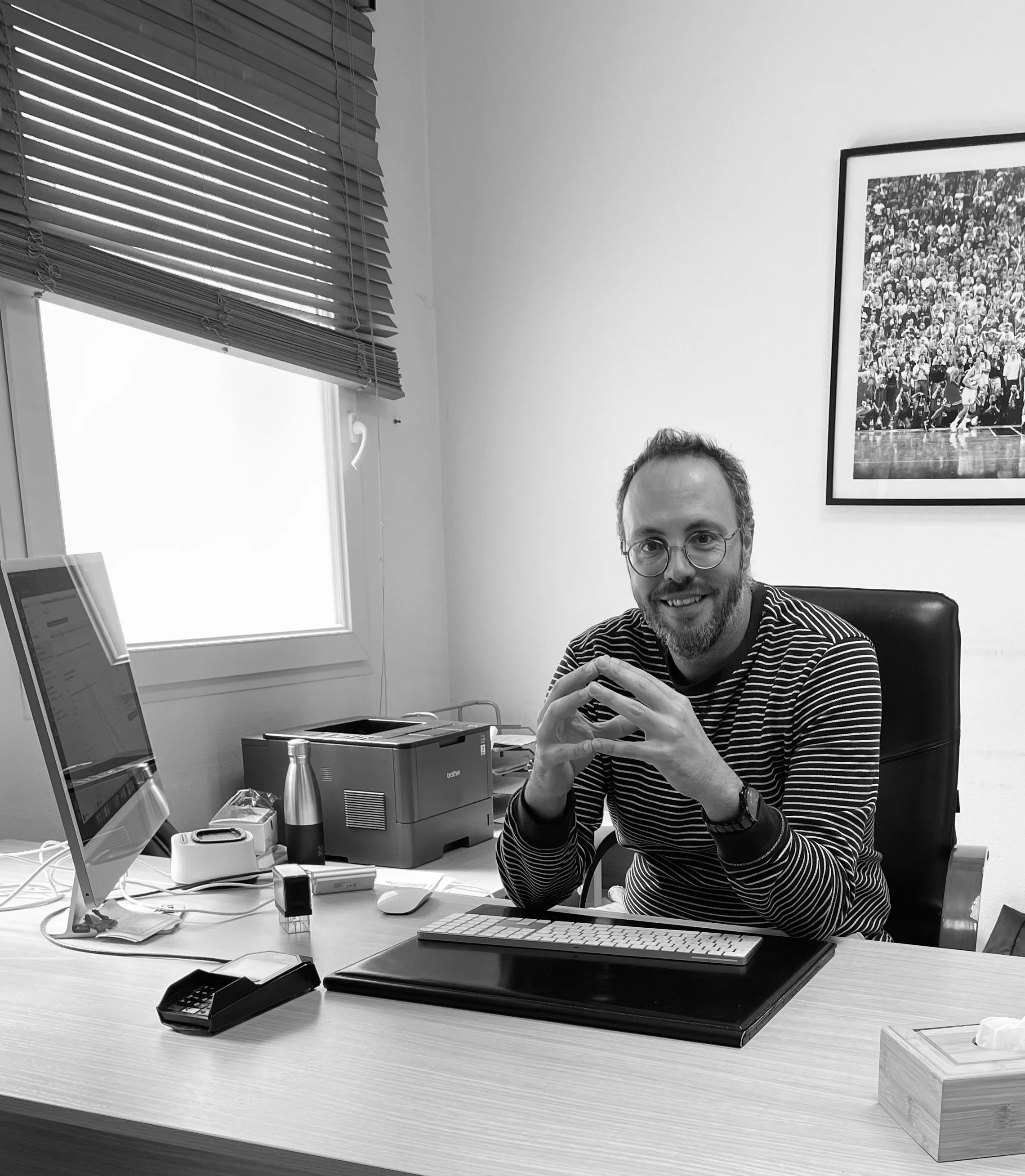 Un homme souriant portant des lunettes, habillé d'une chemise à rayures noires et blanches, assis à un bureau avec un ordinateur, une imprimante et d'autres accessoires.