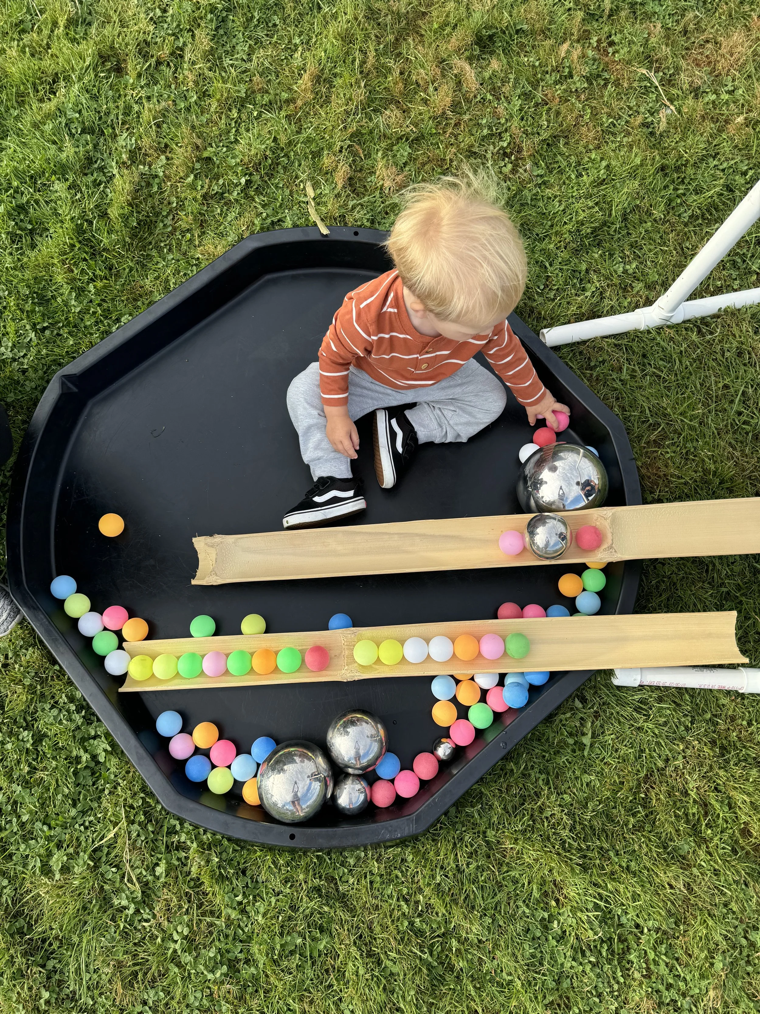 A young child sitting in a black tray with colorful balls and metal bowls, outdoors on grass, playing with balls.
