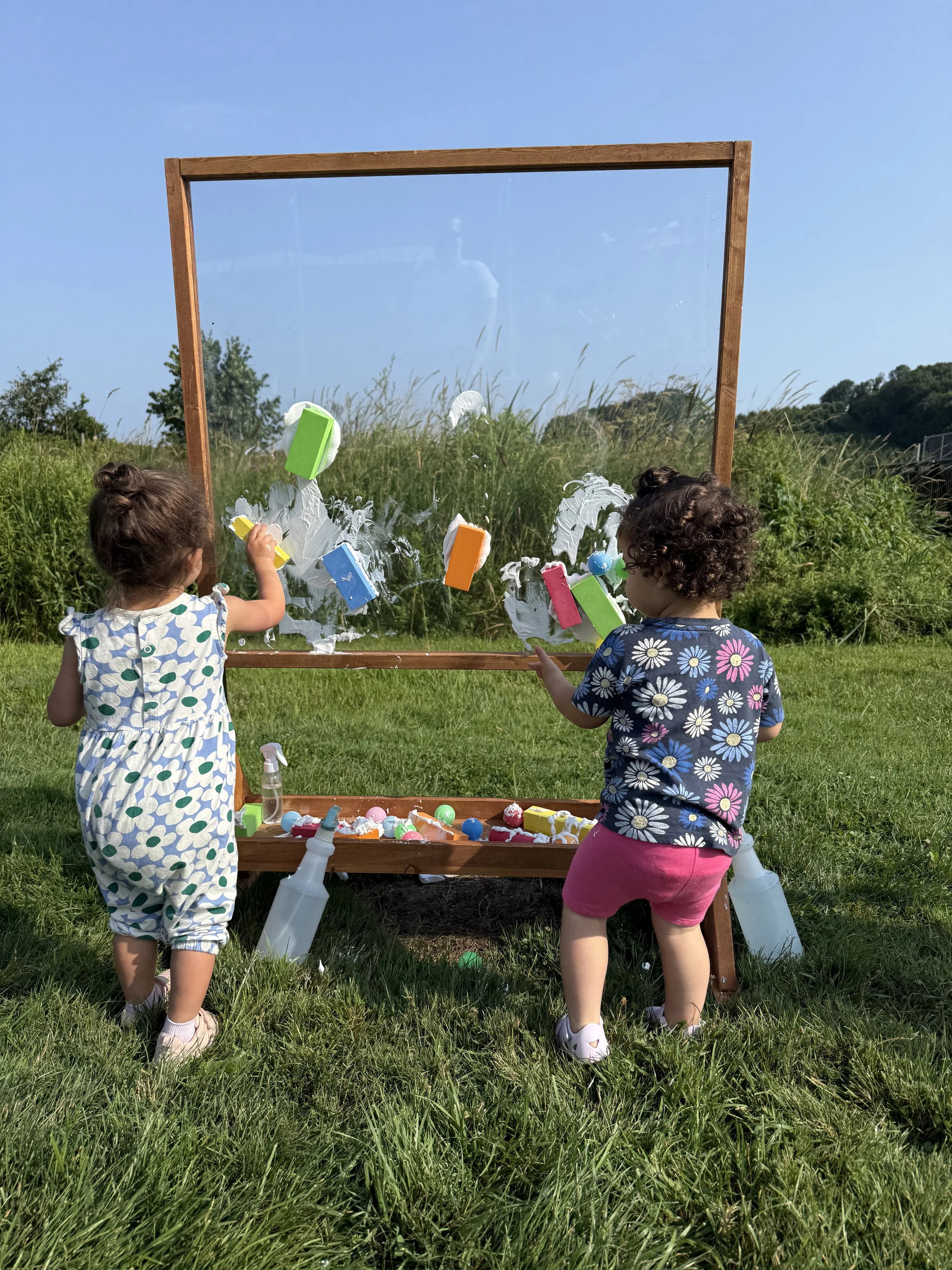 Two young children playing with paint and sponges on a large outdoor mirror.