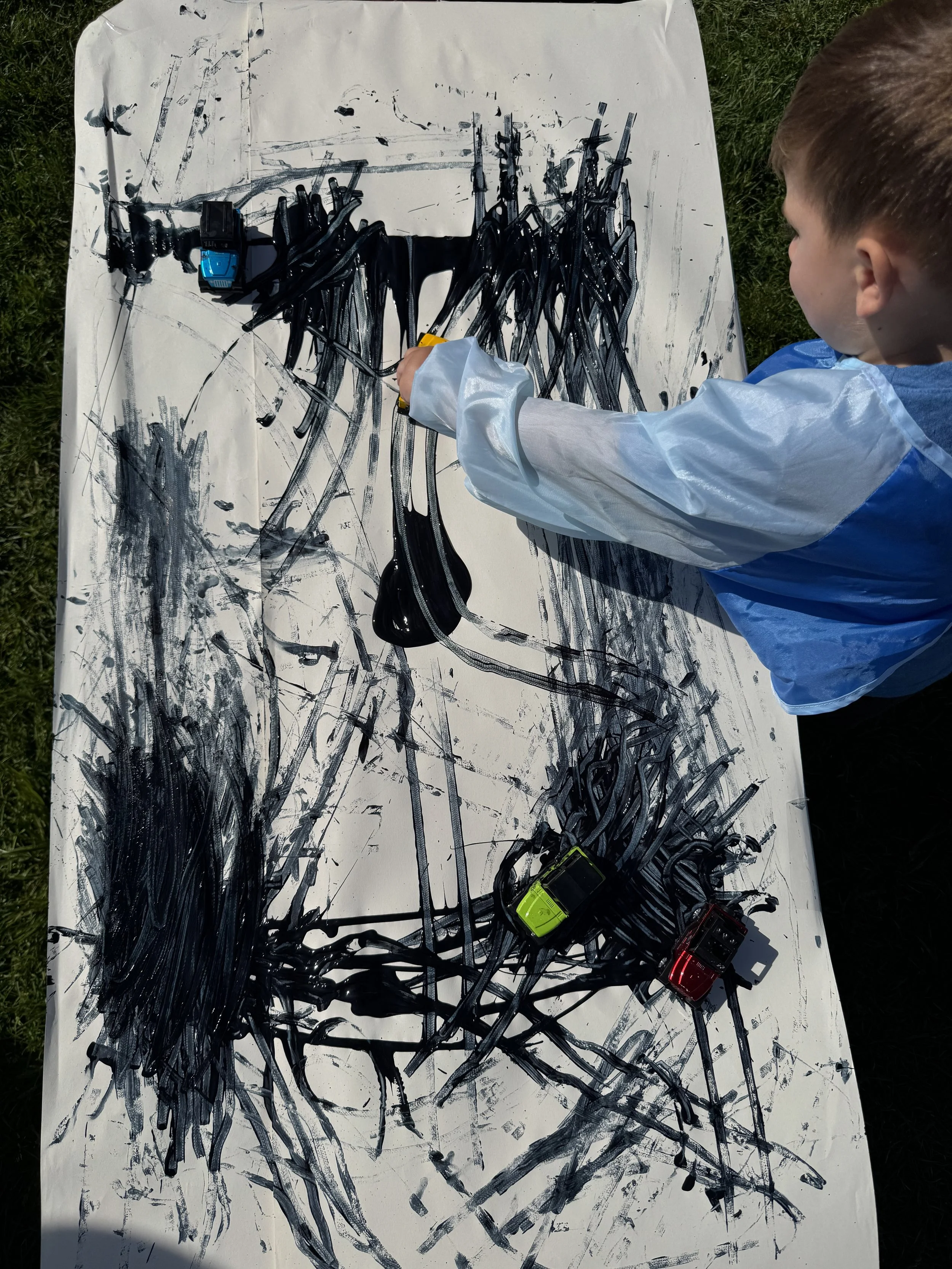 Child painting abstract black lines and streaks on a large white canvas outdoors, with toy cars placed on the canvas.