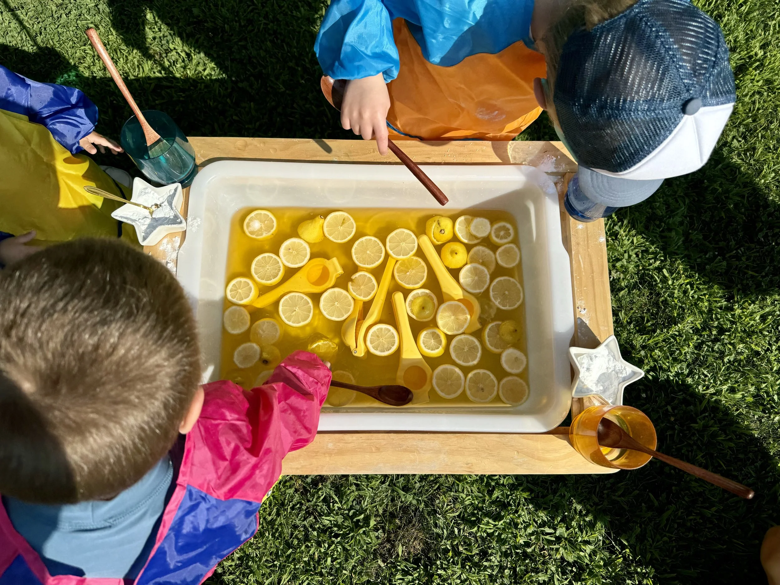 Children making lemonade with sliced lemons in a large white container outdoors.
