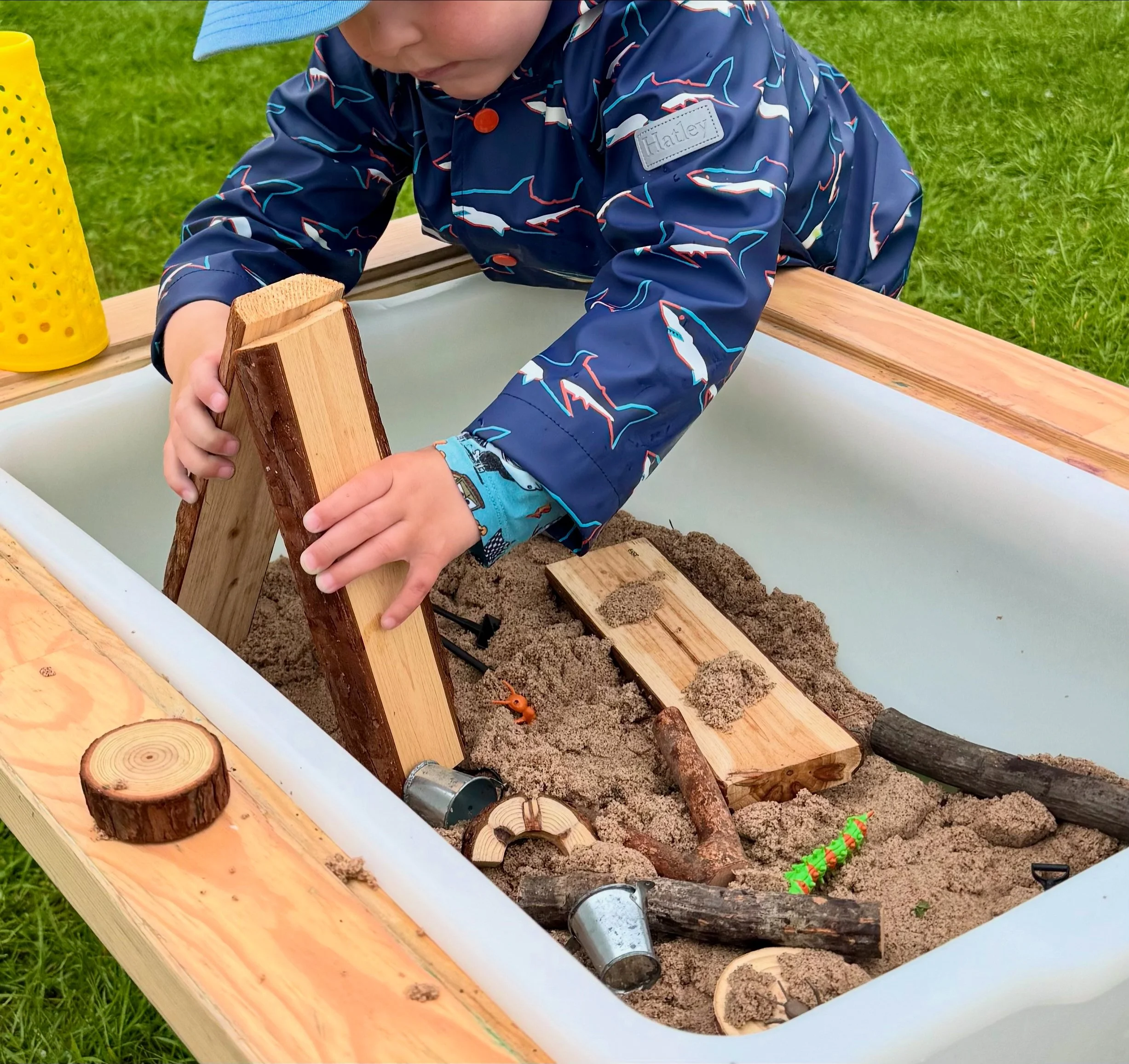 Child playing in sandbox with wooden blocks, sticks, and sand, wearing a navy jacket with shark patterns and a light blue hat.